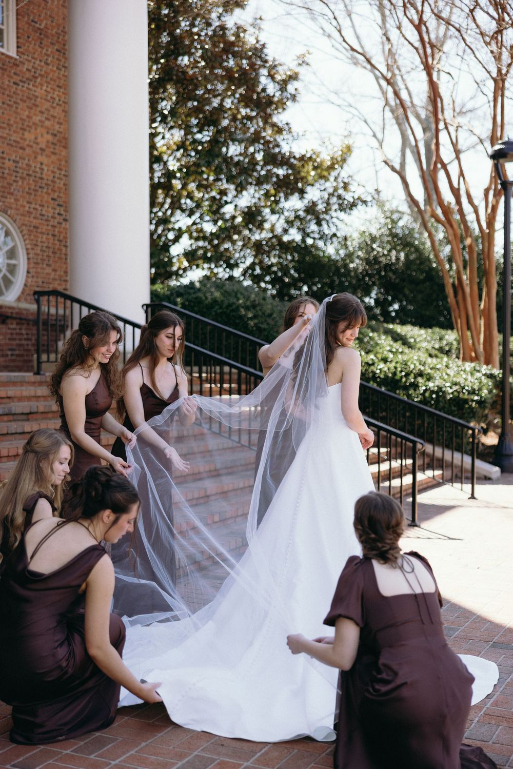 Bridesmaids in burgundy dresses help arrange a long flowing veil on a bride's white wedding gown on brick steps.