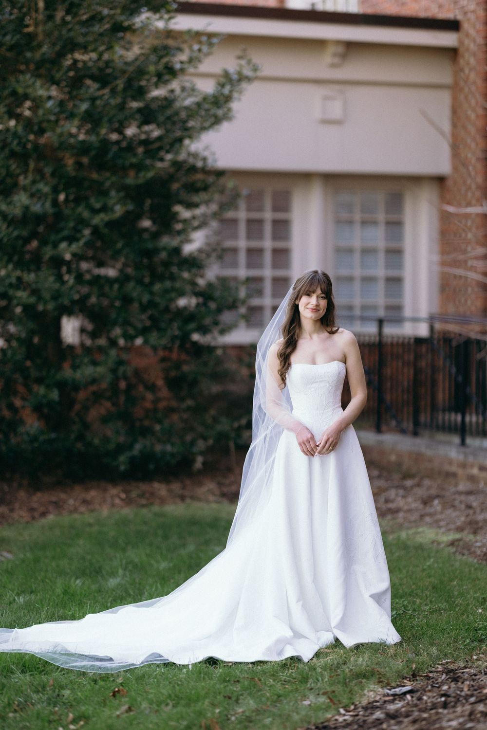 Elegant white wedding gown with sweetheart neckline and flowing train photographed on grassy lawn by brick building.