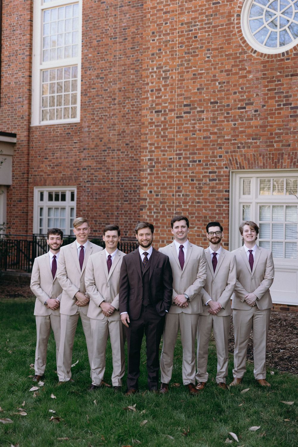A group of groomsmen in tan suits pose together against a brick building exterior.