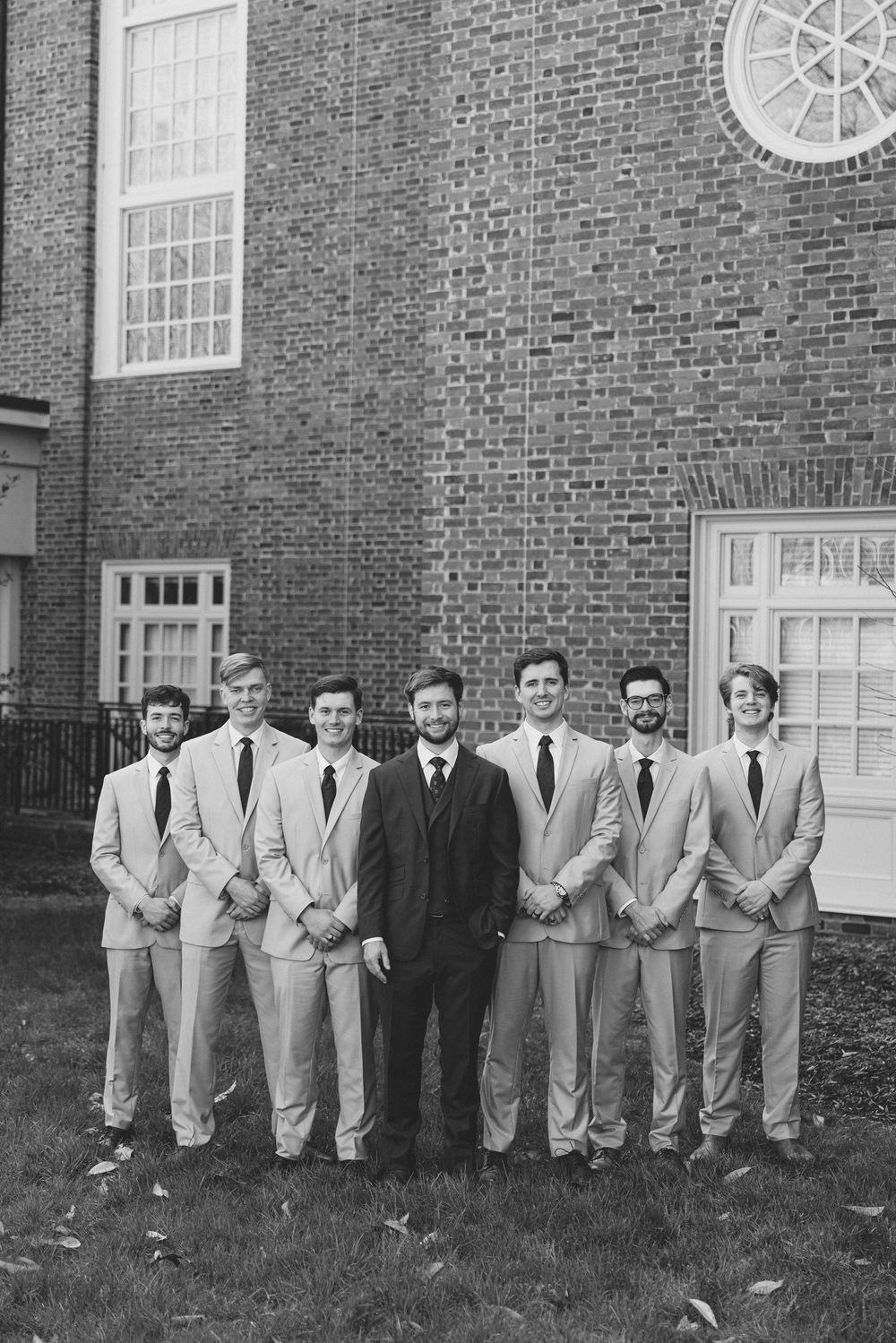 A group of groomsmen in light colored suits pose with one person in a dark suit against a brick building exterior.