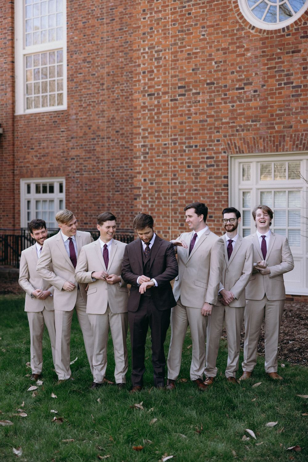 A line of groomsmen in tan suits pose playfully against a brick building wall.
