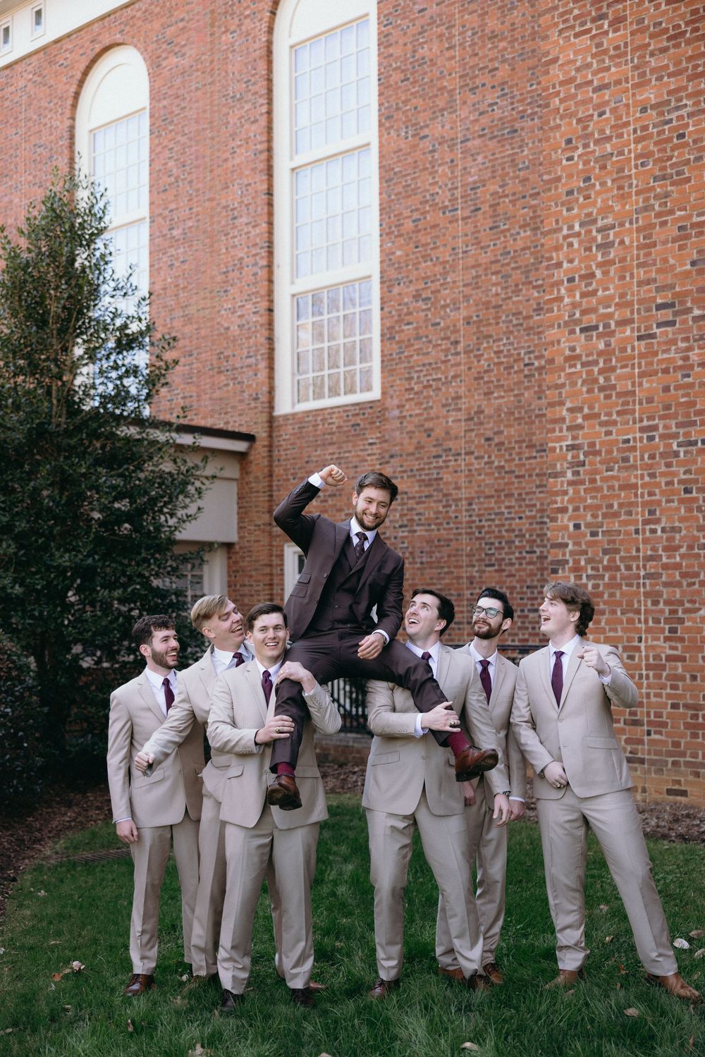 Wedding party members in beige suits strike playful poses outside a brick church building.