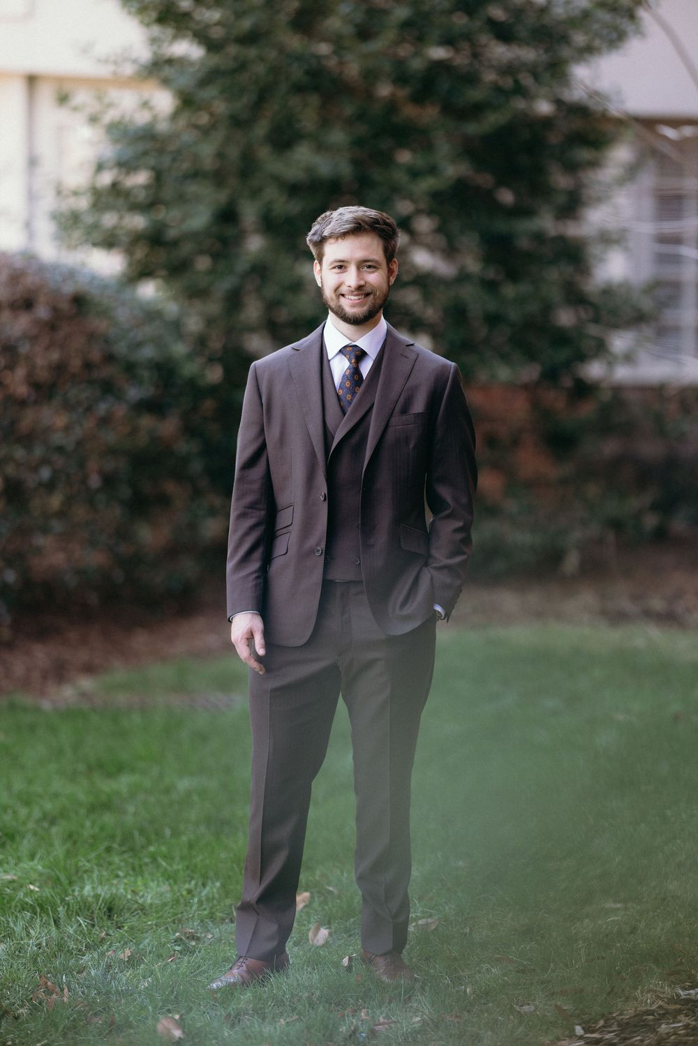 A person in a dark brown three-piece suit stands on grass with trees in the background.