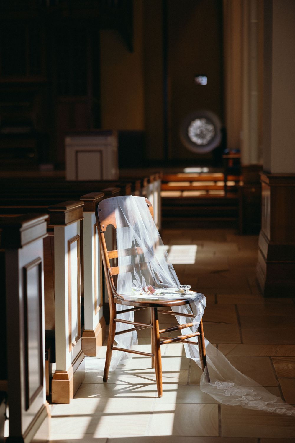 A wedding veil draped over a wooden chair in a sunlit church aisle creates an ethereal scene.