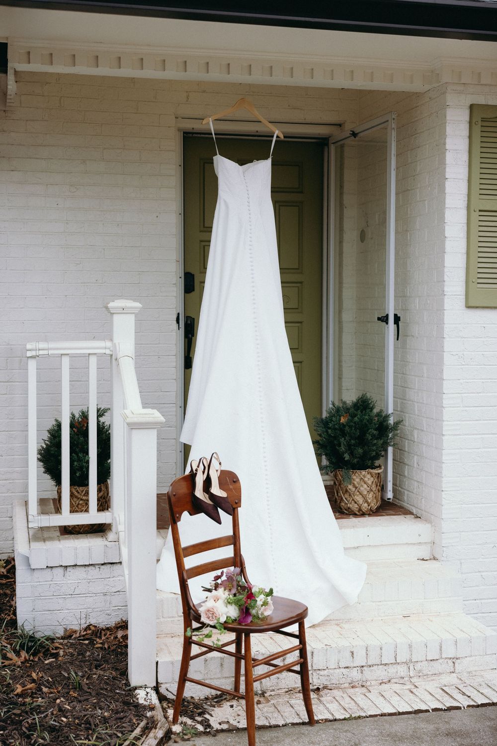 White wedding dresses hang elegantly from wooden chairs on a classic southern porch with white brick columns.