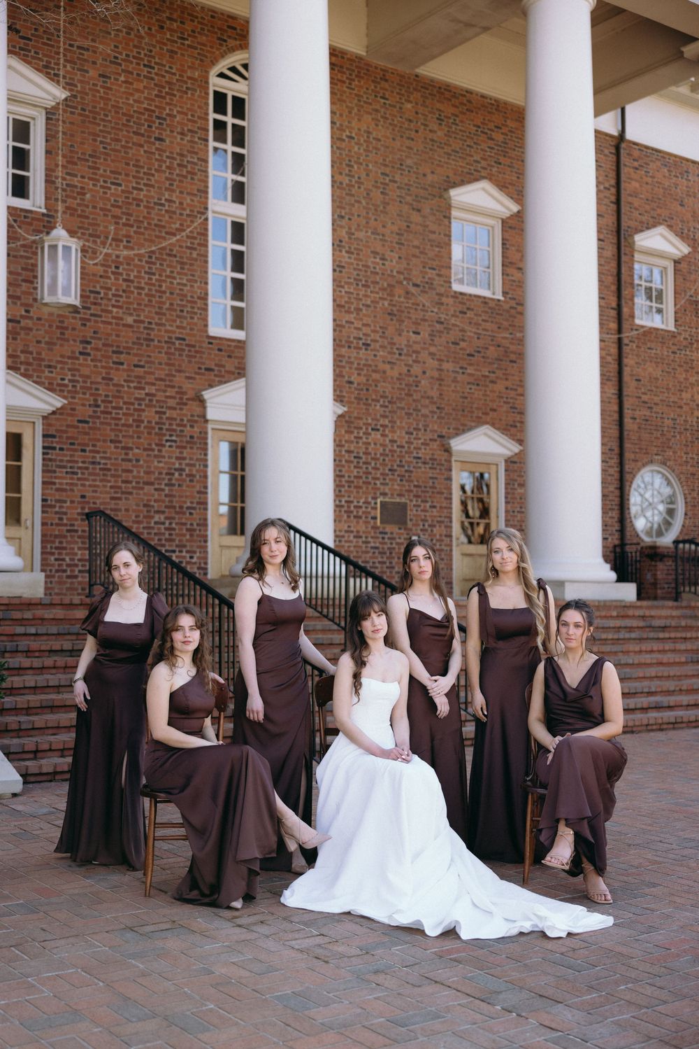 A wedding party poses on brick steps with white columns, featuring bridesmaids in brown dresses and a bride in white.