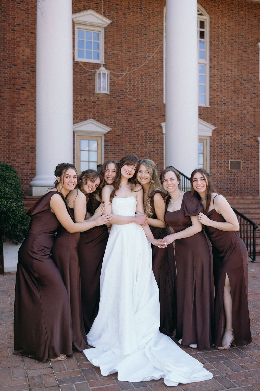 Wedding party poses in front of brick building with white columns, bridesmaids wearing brown dresses.