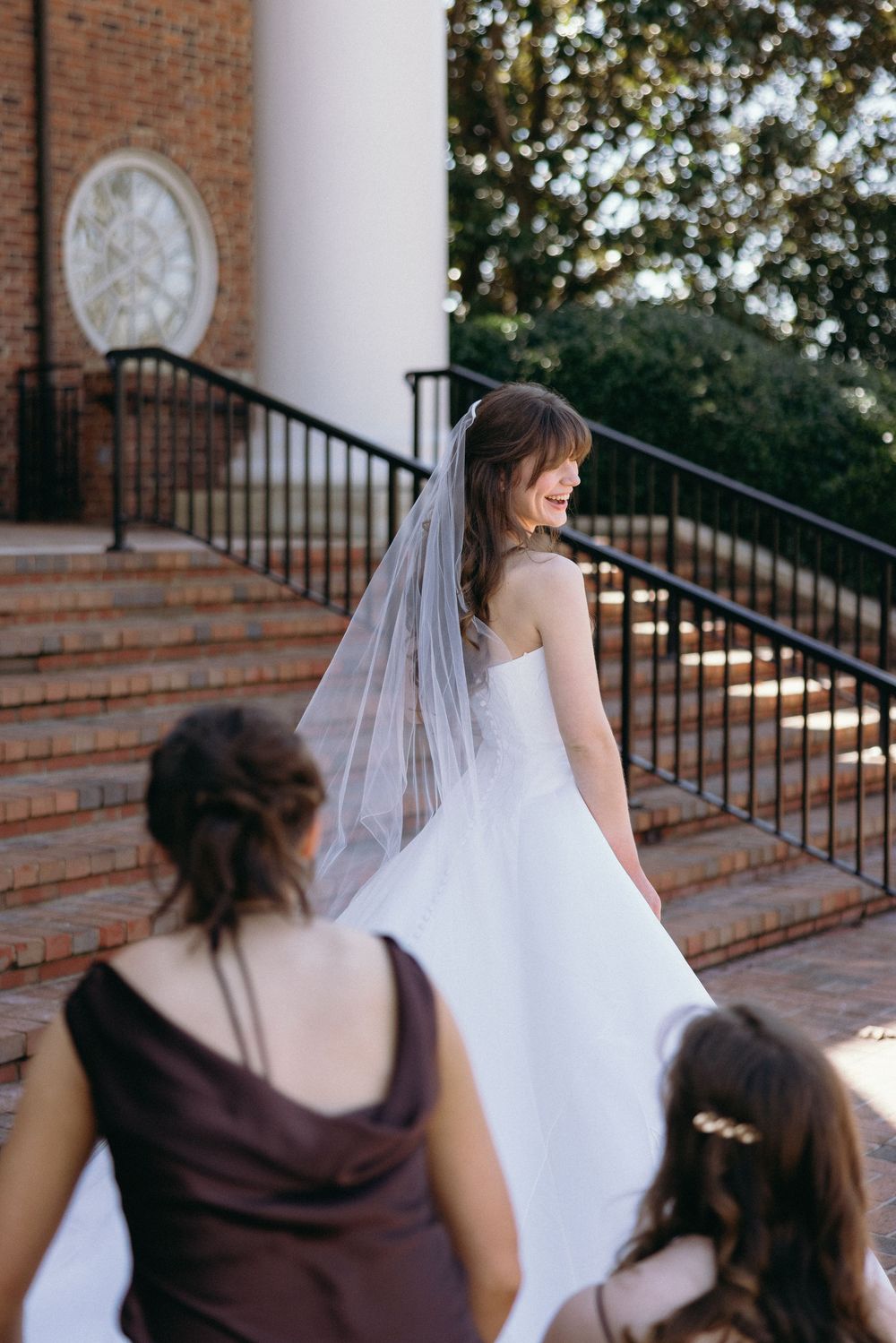 A bride in a white wedding gown stands on brick church steps while bridesmaids in burgundy dresses look on from below.