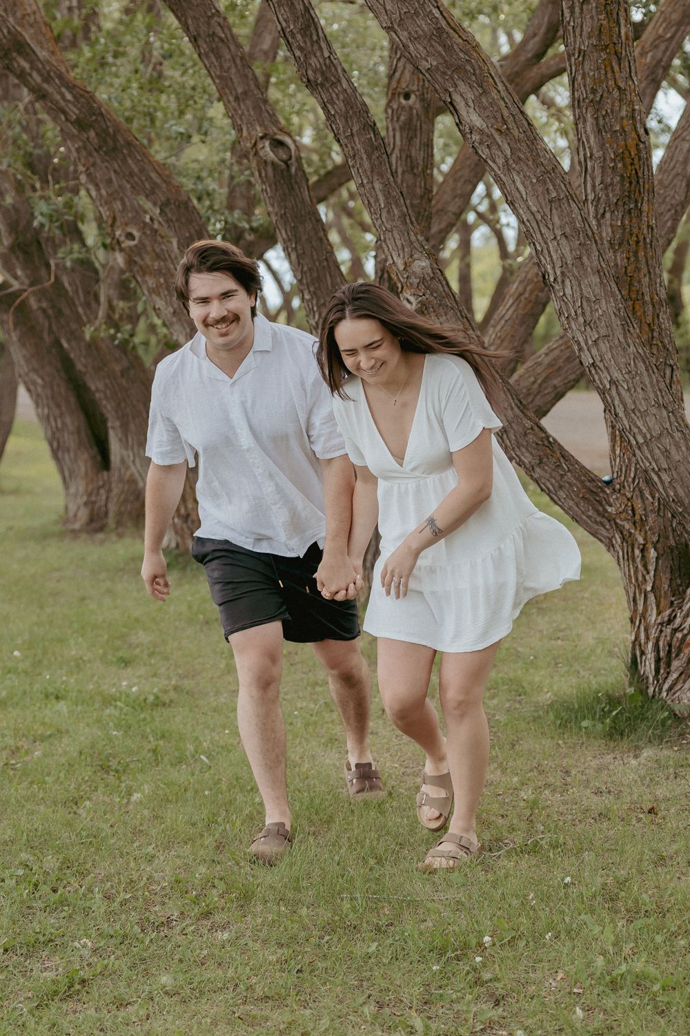 Playful couple in white outfits running through grove of trees.