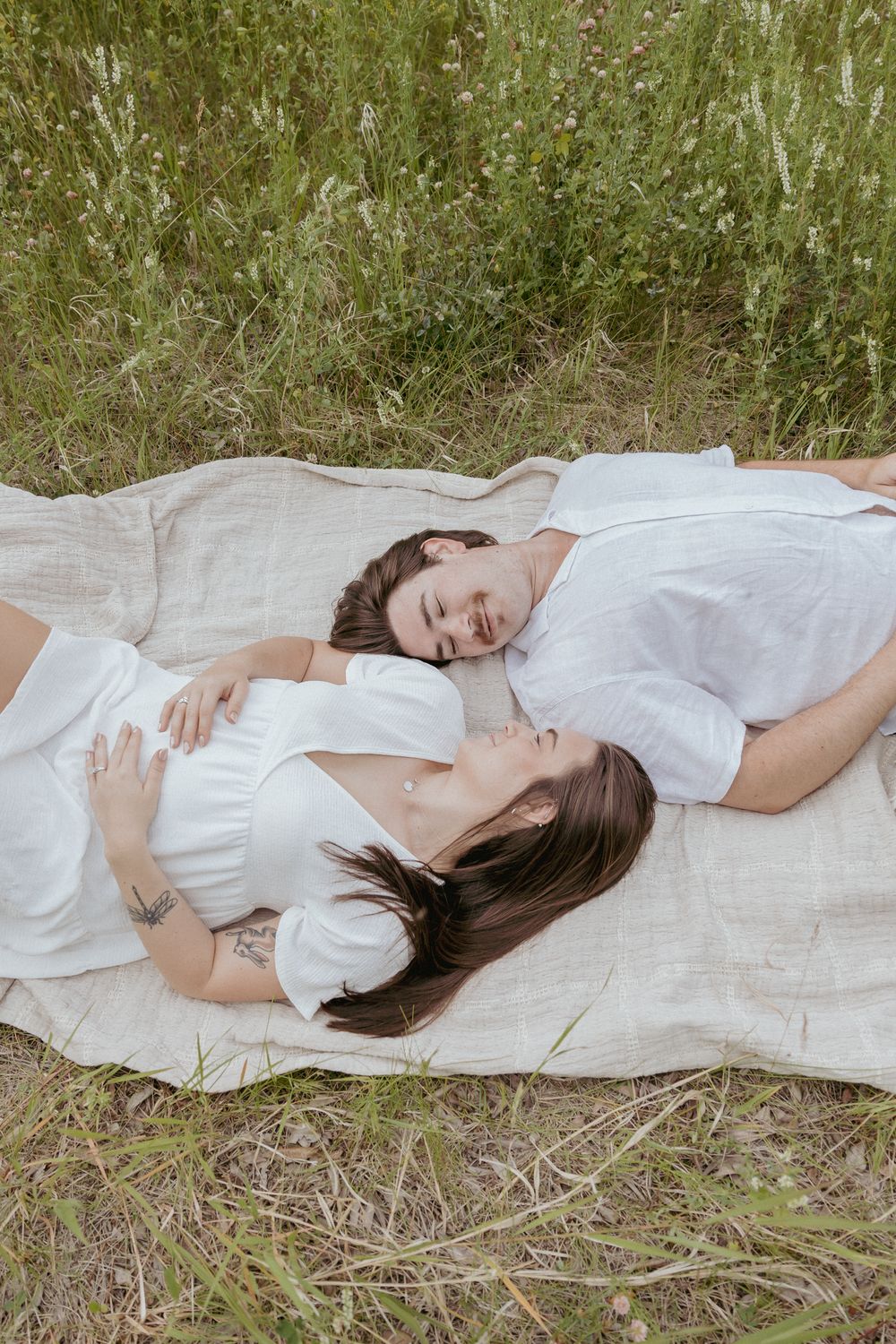 A couple in white clothing resting peacefully on a blanket in a grassy field.