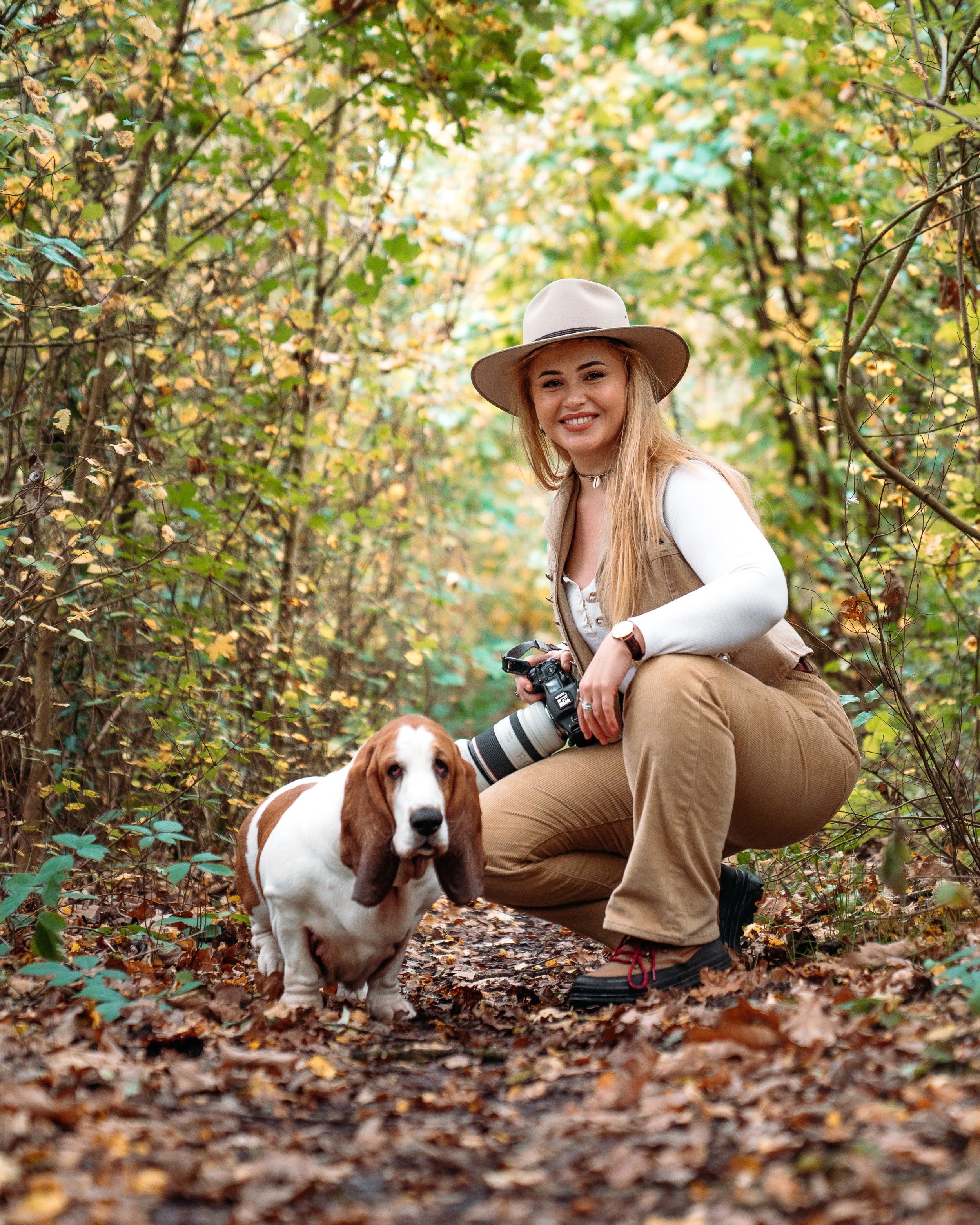Photographer with camera kneeling beside Basset Hound on autumn forest trail.