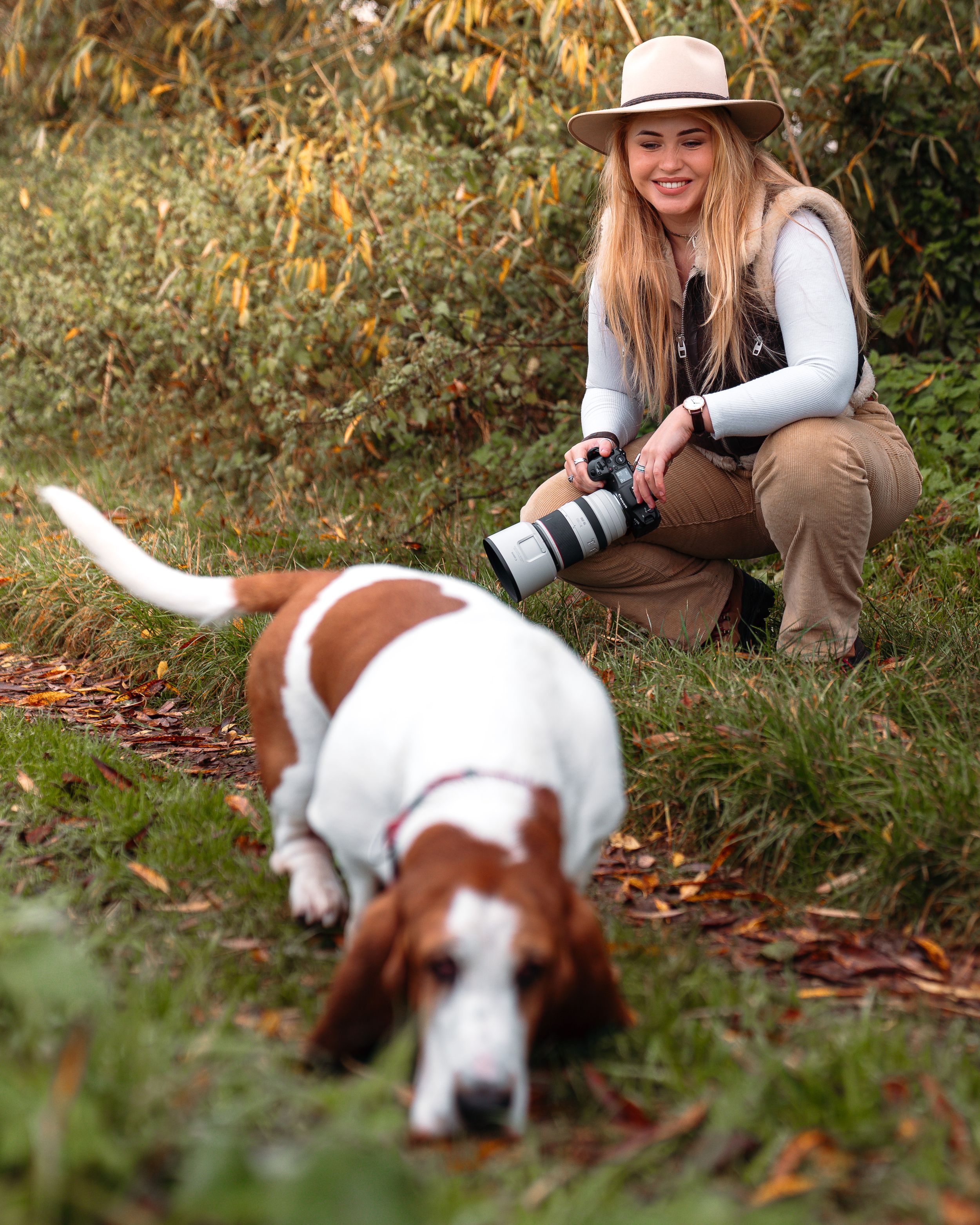 Photographer in straw hat crouching with camera while brown and white dog explores fallen leaves.