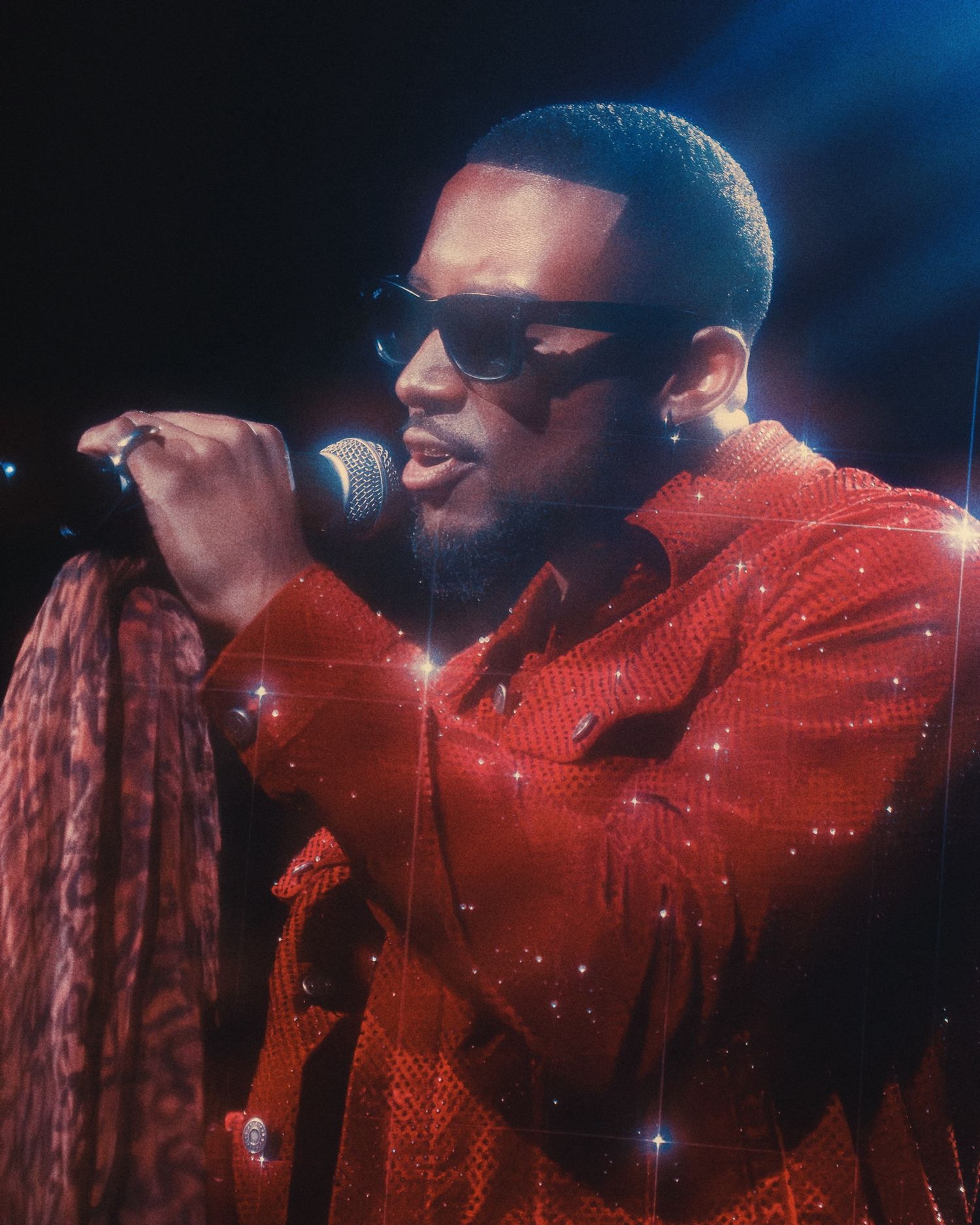A performer in a sparkling red outfit sings into a microphone against a dark blue background during a stage performance.