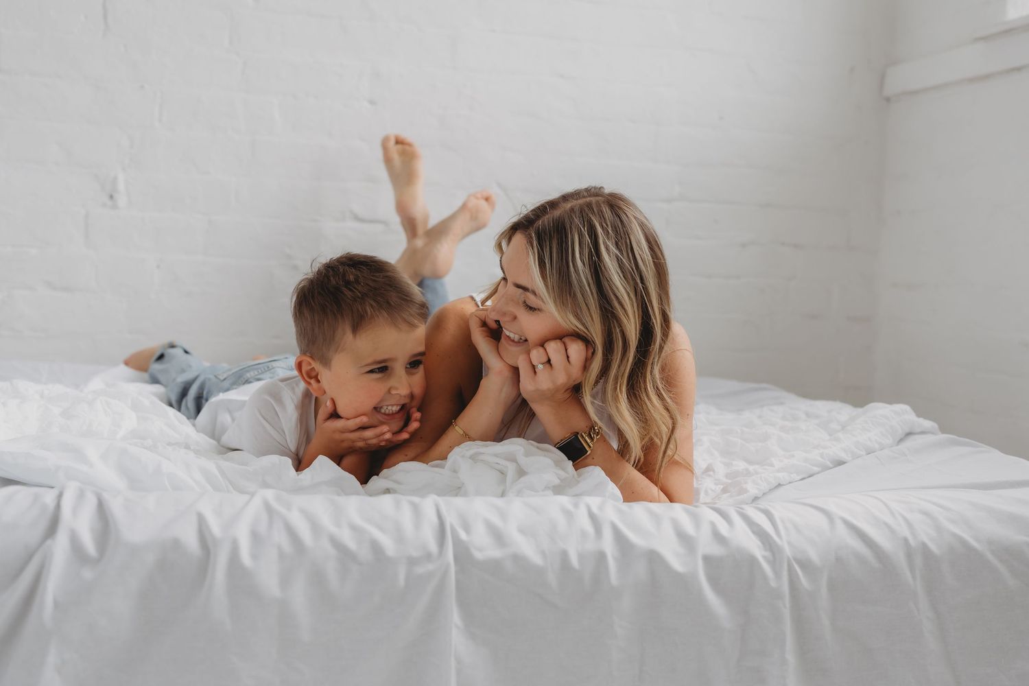 Parent and child share a playful moment while relaxing together on white bedding in a bright room.
