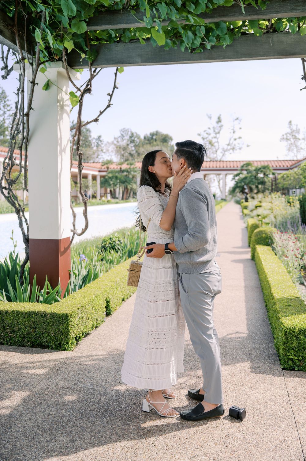 A romantic couple embraces in a garden pathway surrounded by manicured hedges and white columns.
