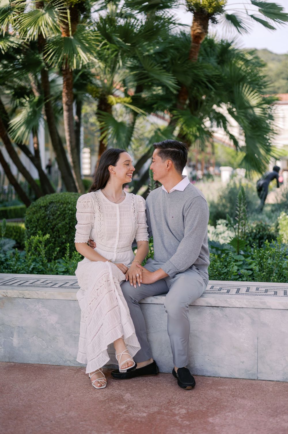 A couple sits together on a concrete ledge surrounded by palm trees in coordinating white and gray outfits.