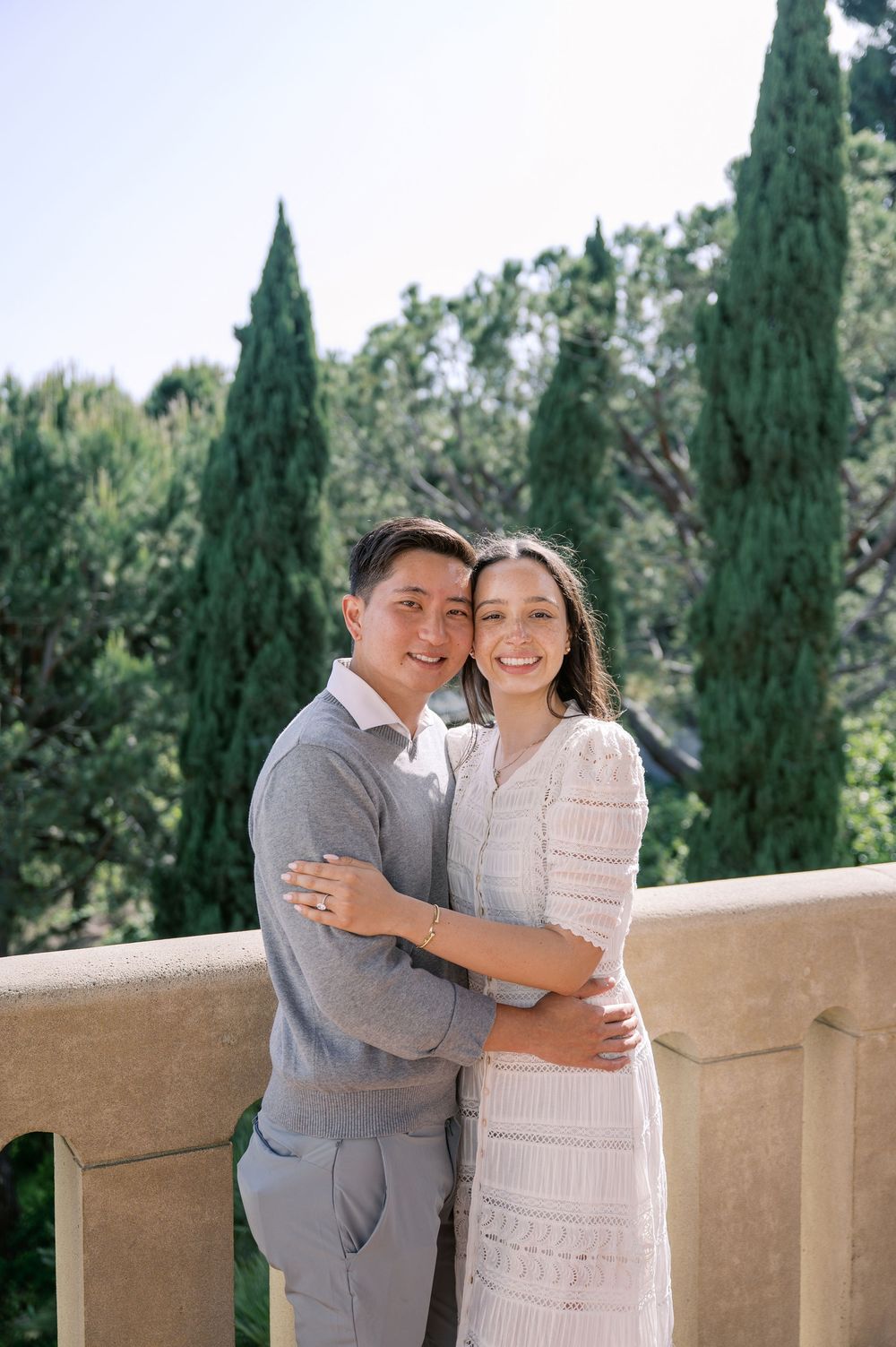 A couple in gray and white attire embrace while standing on a terrace with cypress trees in the background.