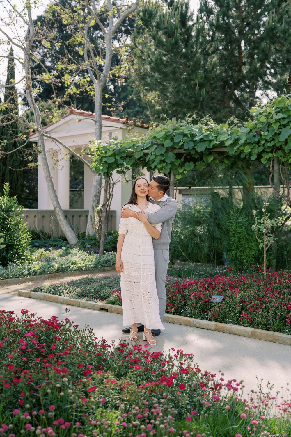 A couple shares a romantic moment in a garden path surrounded by vibrant pink flowers and lush greenery.