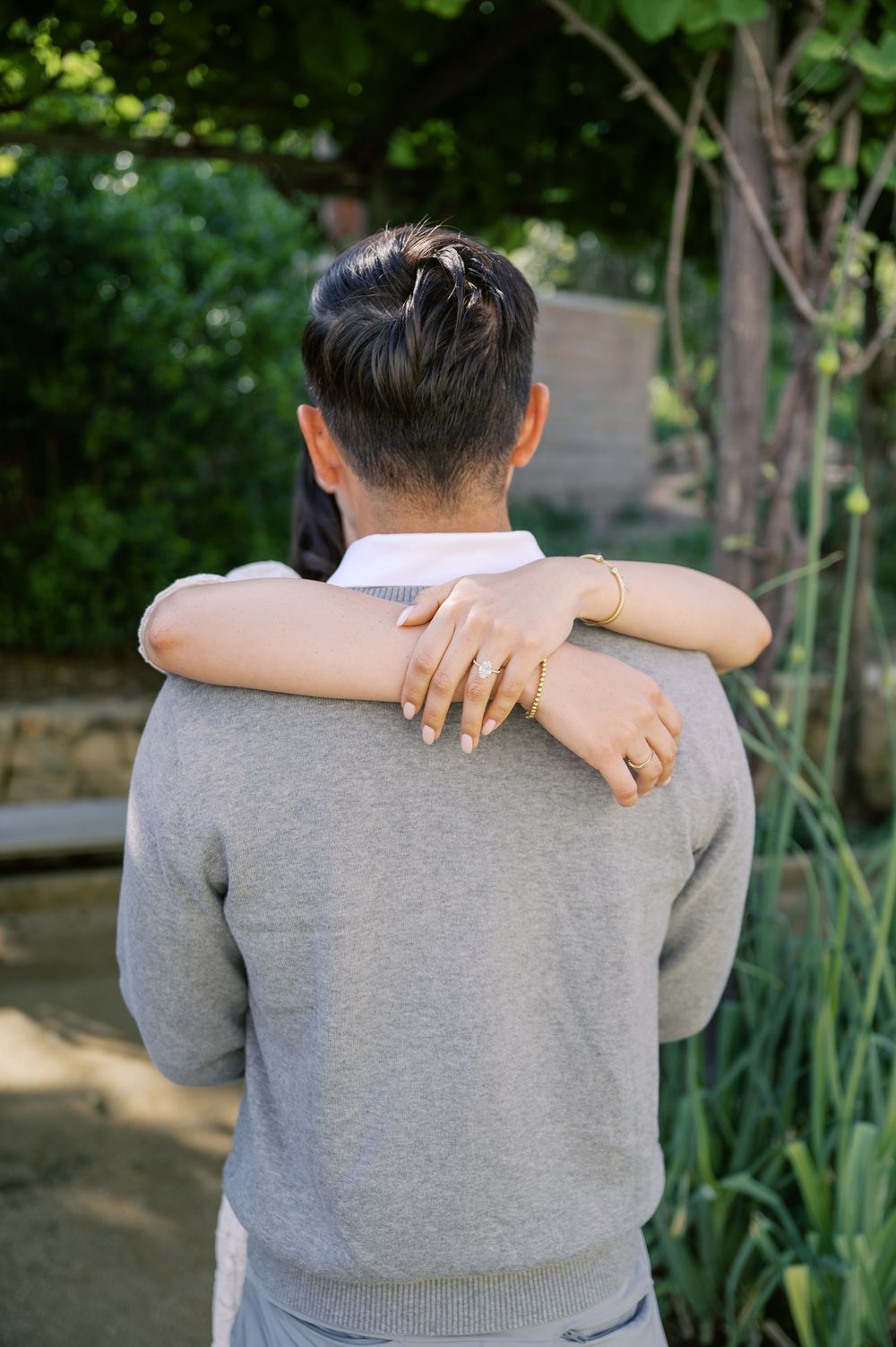 A person in a gray sweater is photographed from behind with their arms crossed against a green garden background.