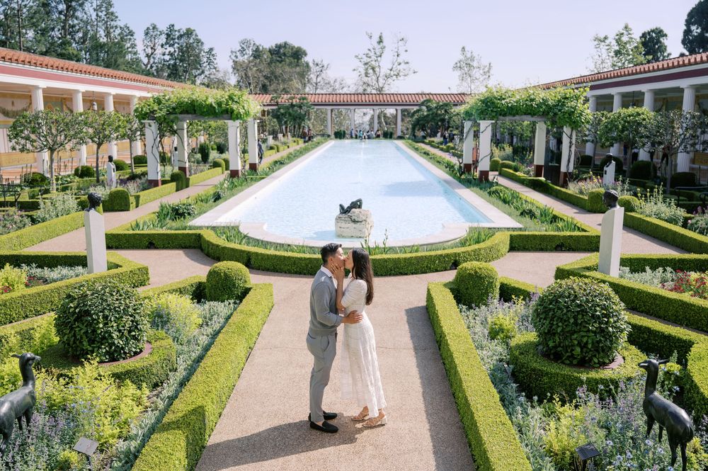 A couple embraces in a formal garden with manicured hedges and a large fountain at the Getty Villa in Los Angeles.