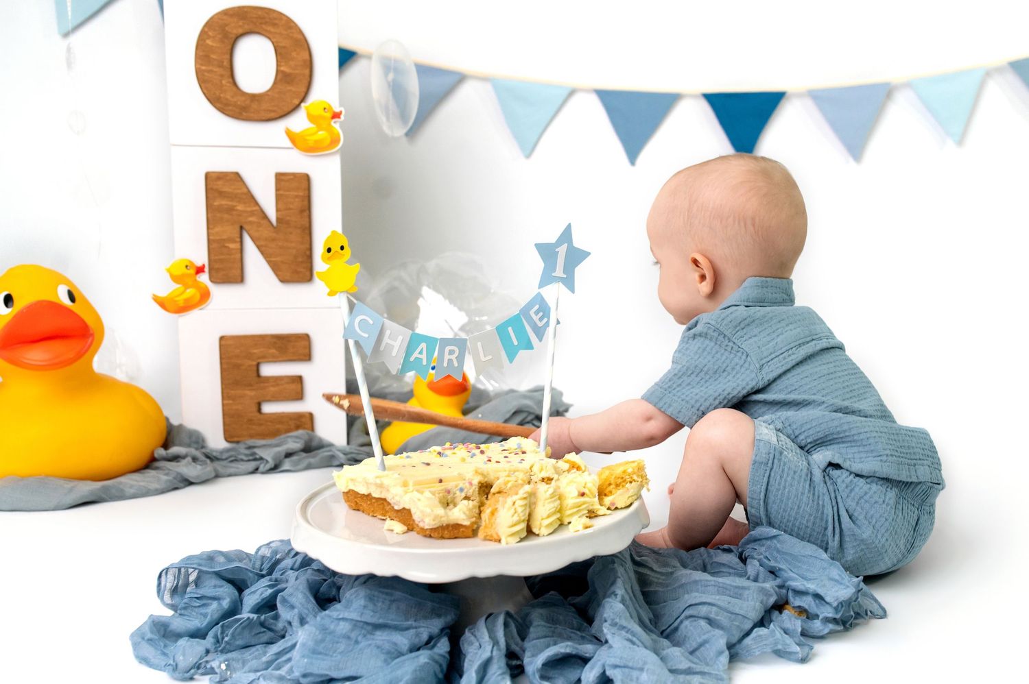 Baby wearing blue outfit lies on white surface while playing with yellow rubber duck toy during milestone photoshoot.
