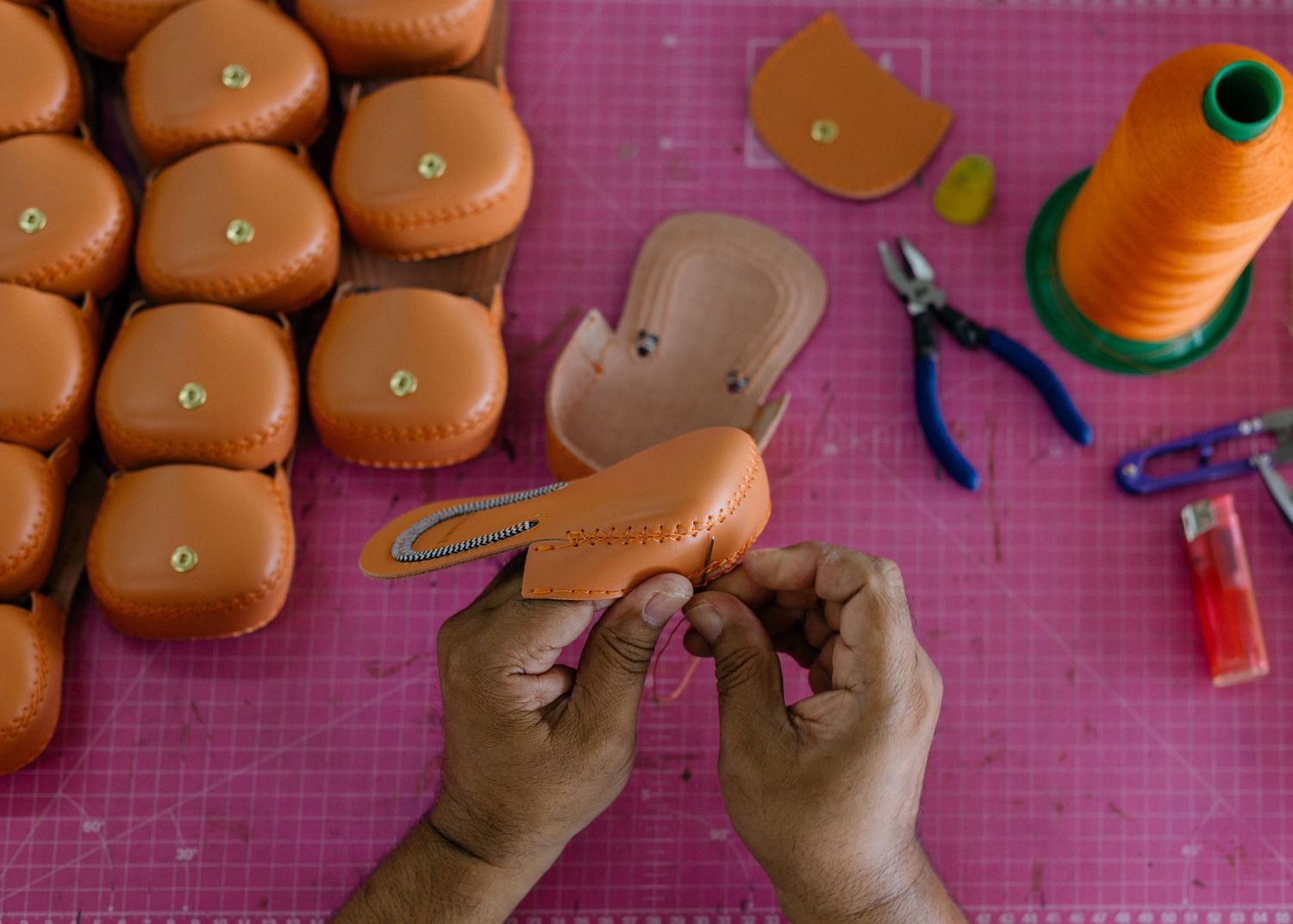 Hands working with leather crafting materials on a pink surface with sewing tools.