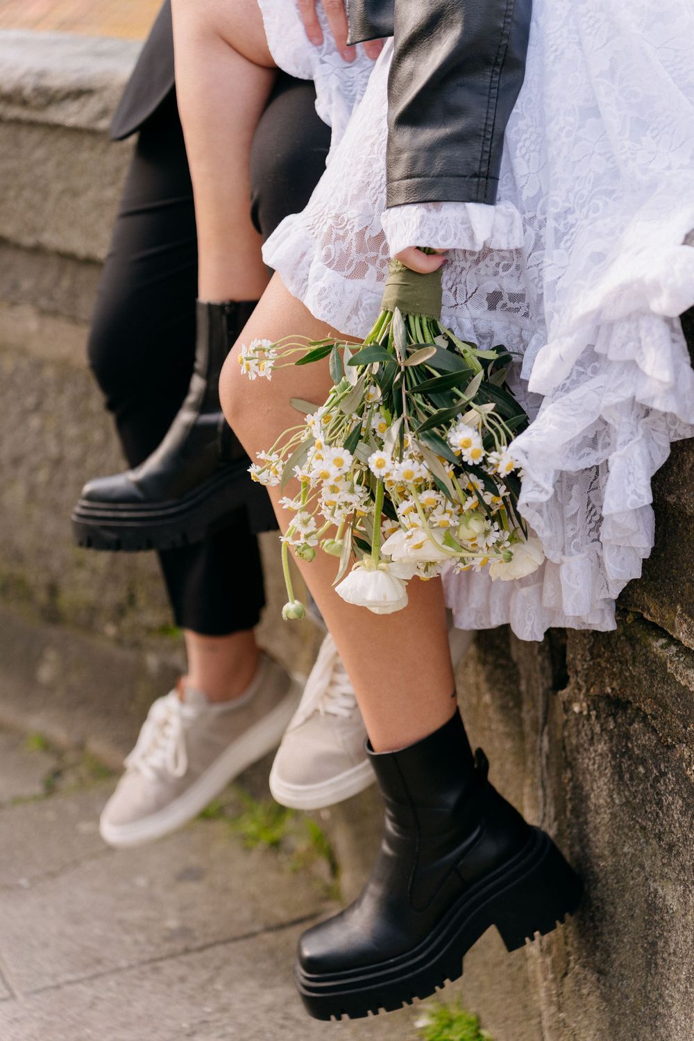 People wearing black boots and white dresses sitting on a stone wall with flowers.