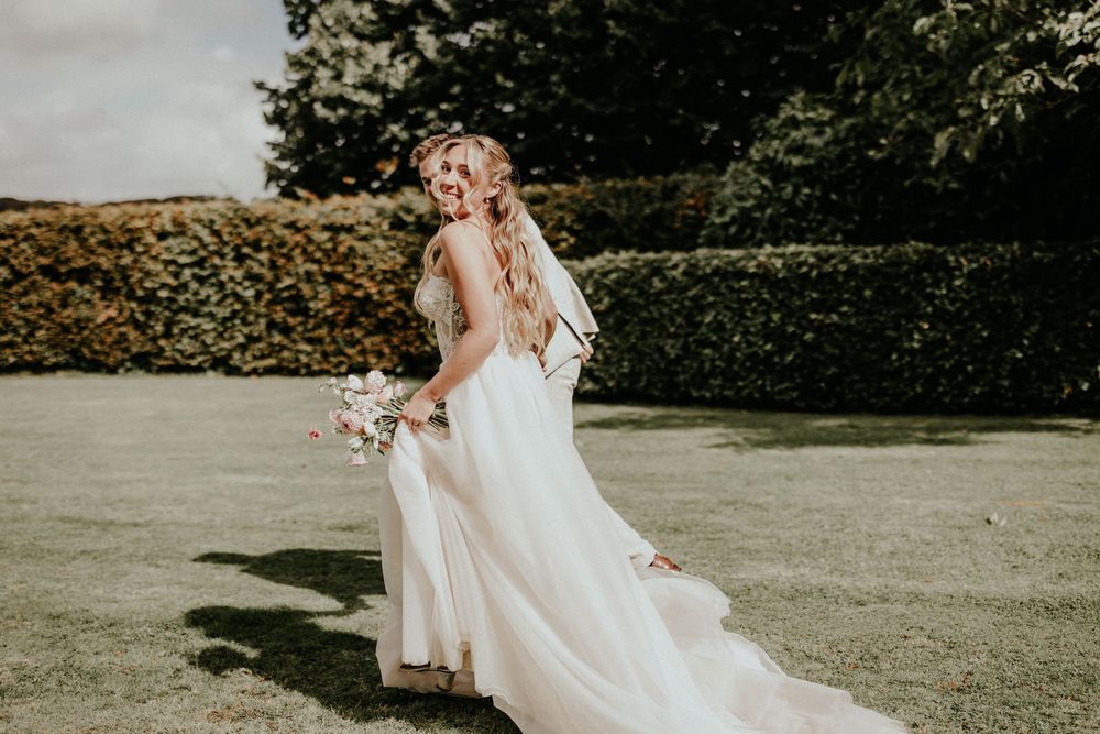 A bride in a flowing white dress spins joyfully on a grassy lawn during a sunny outdoor wedding celebration.