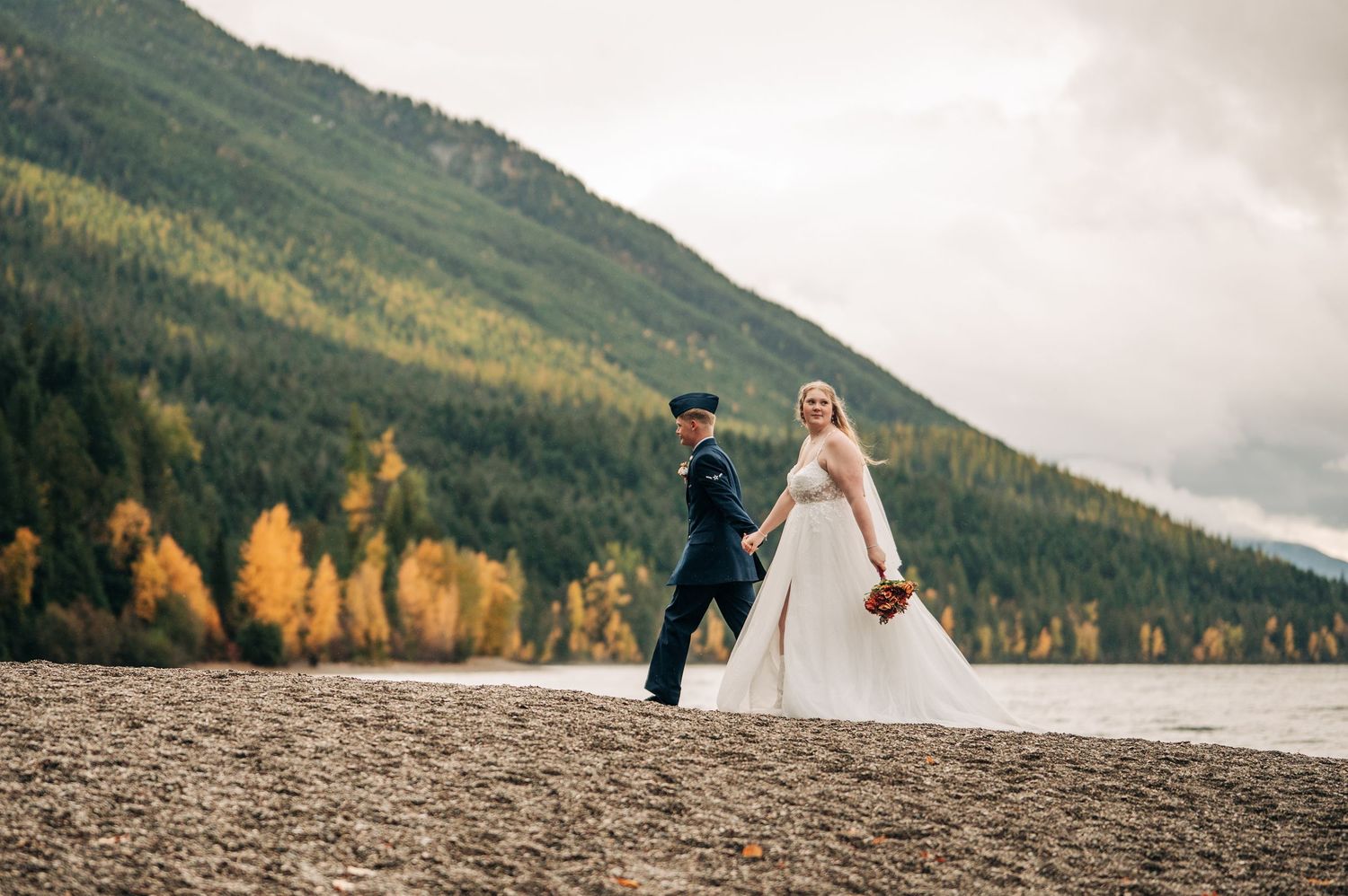 A couple in wedding attire walks along a scenic mountain lake shore with golden autumn trees in the background.