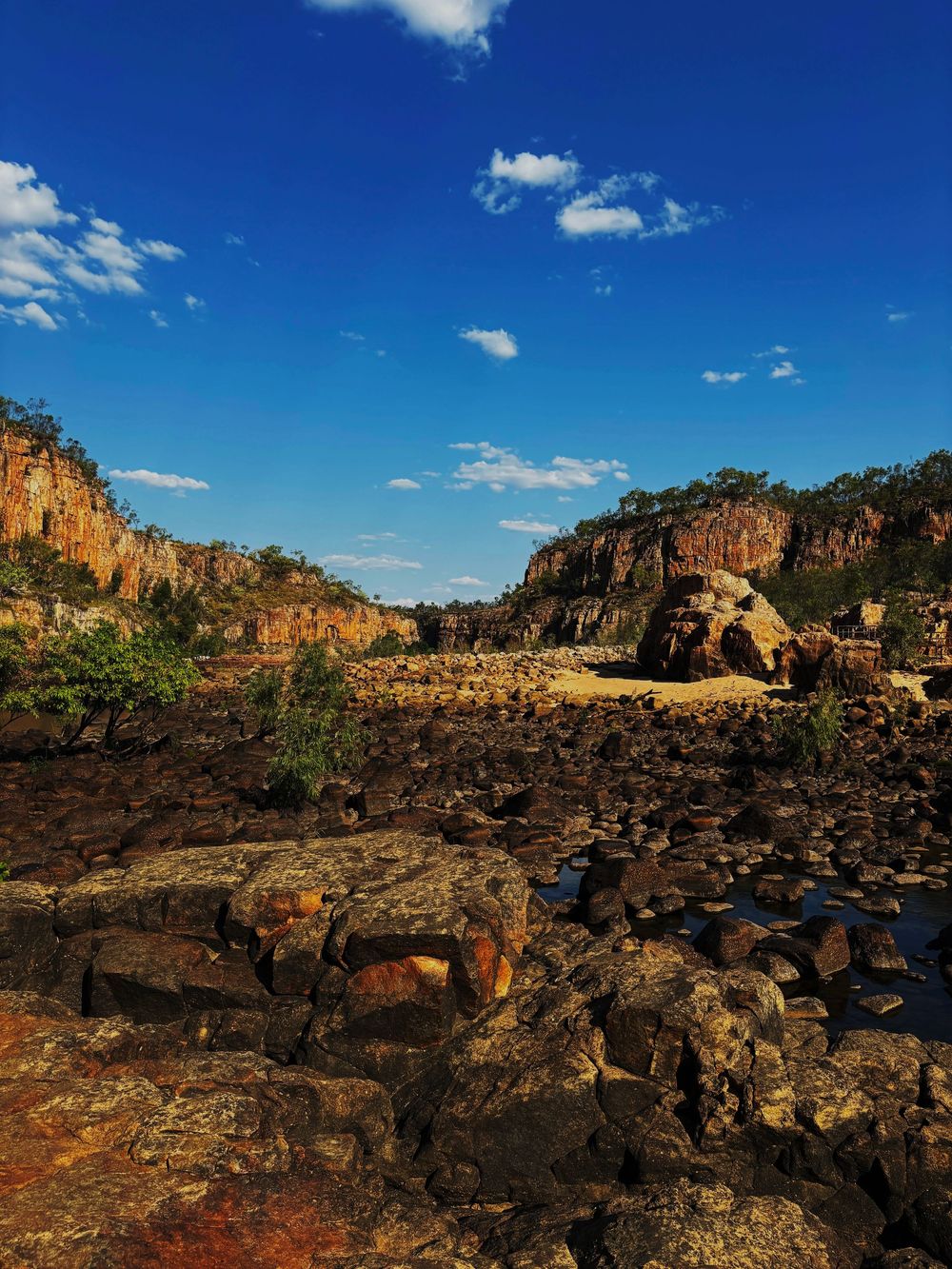 Rocky landscape with red cliffs, scattered rocks, and greenery under a vivid blue sky in Australian outback.