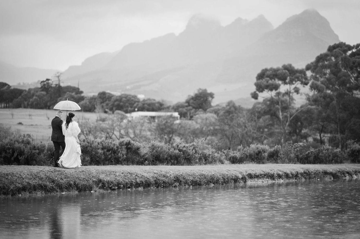 Couple sharing umbrella in rain near reflective pond with misty mountain backdrop in black and white wedding photo.