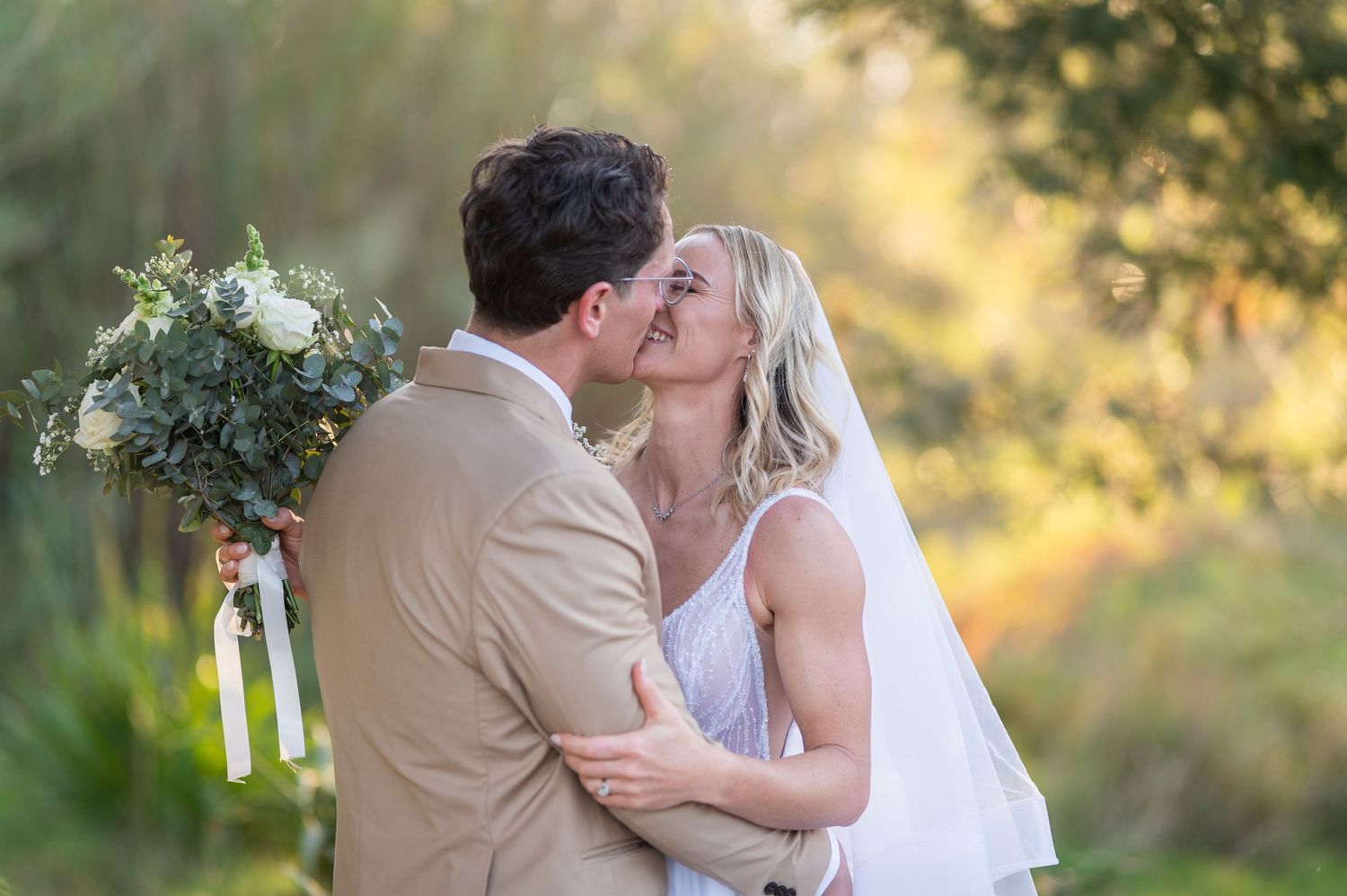 Bride and groom share a romantic kiss outdoors, bride holding white floral bouquet with greenery, wearing veil and white dress.