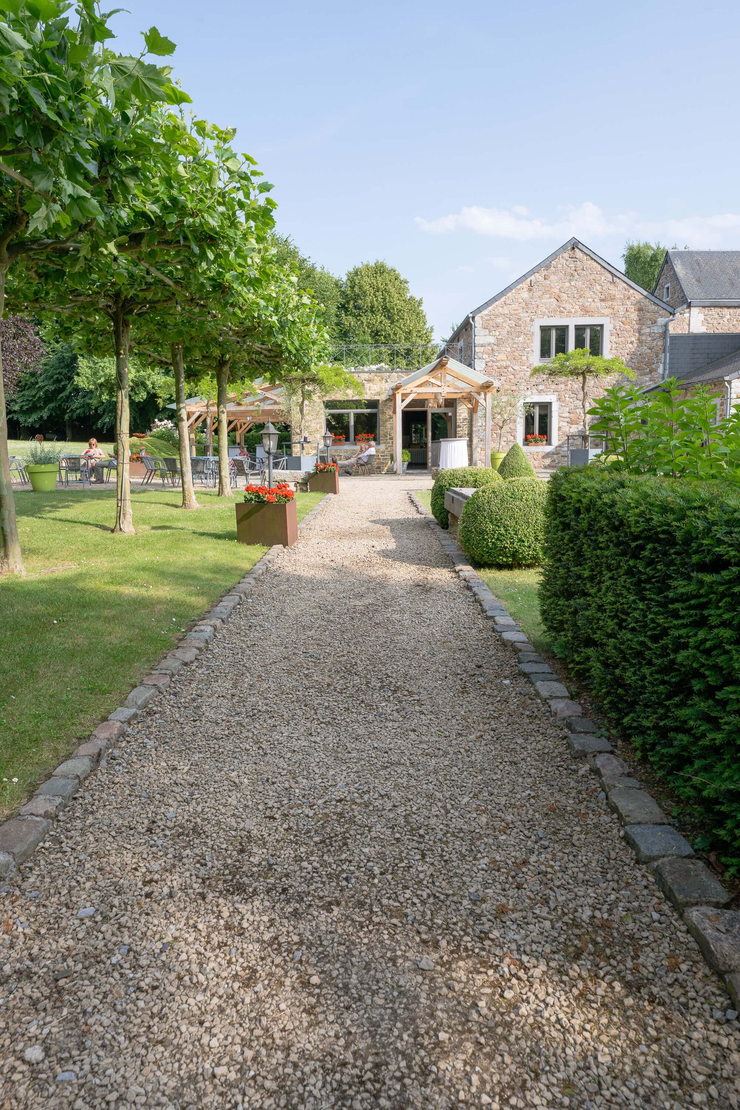 A gravel path lined with trees and hedges leads to a rustic stone house with a welcoming entrance.
