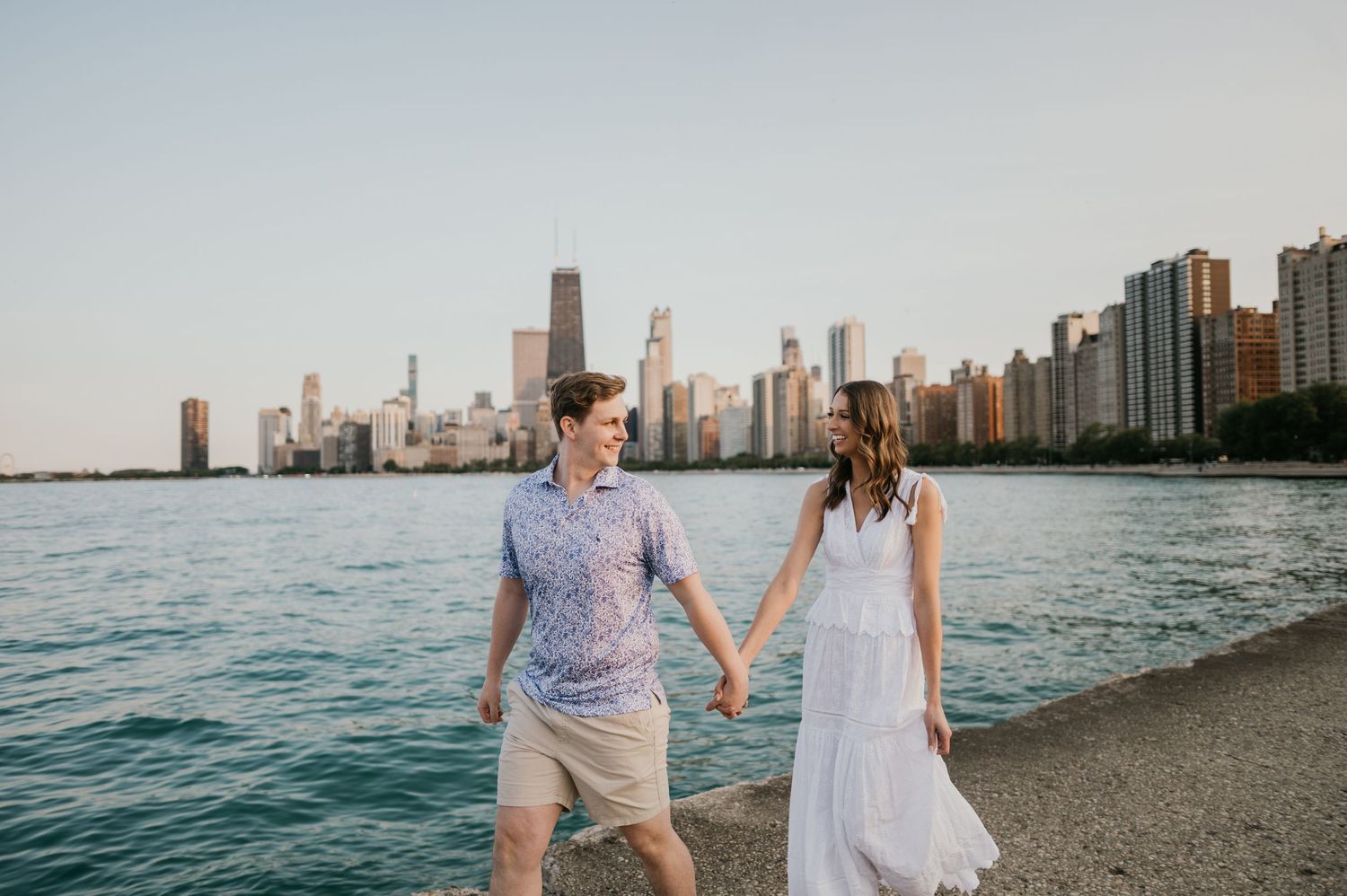 Couple walking hand in hand along Lake Michigan shore with Chicago skyline in background during sunset.