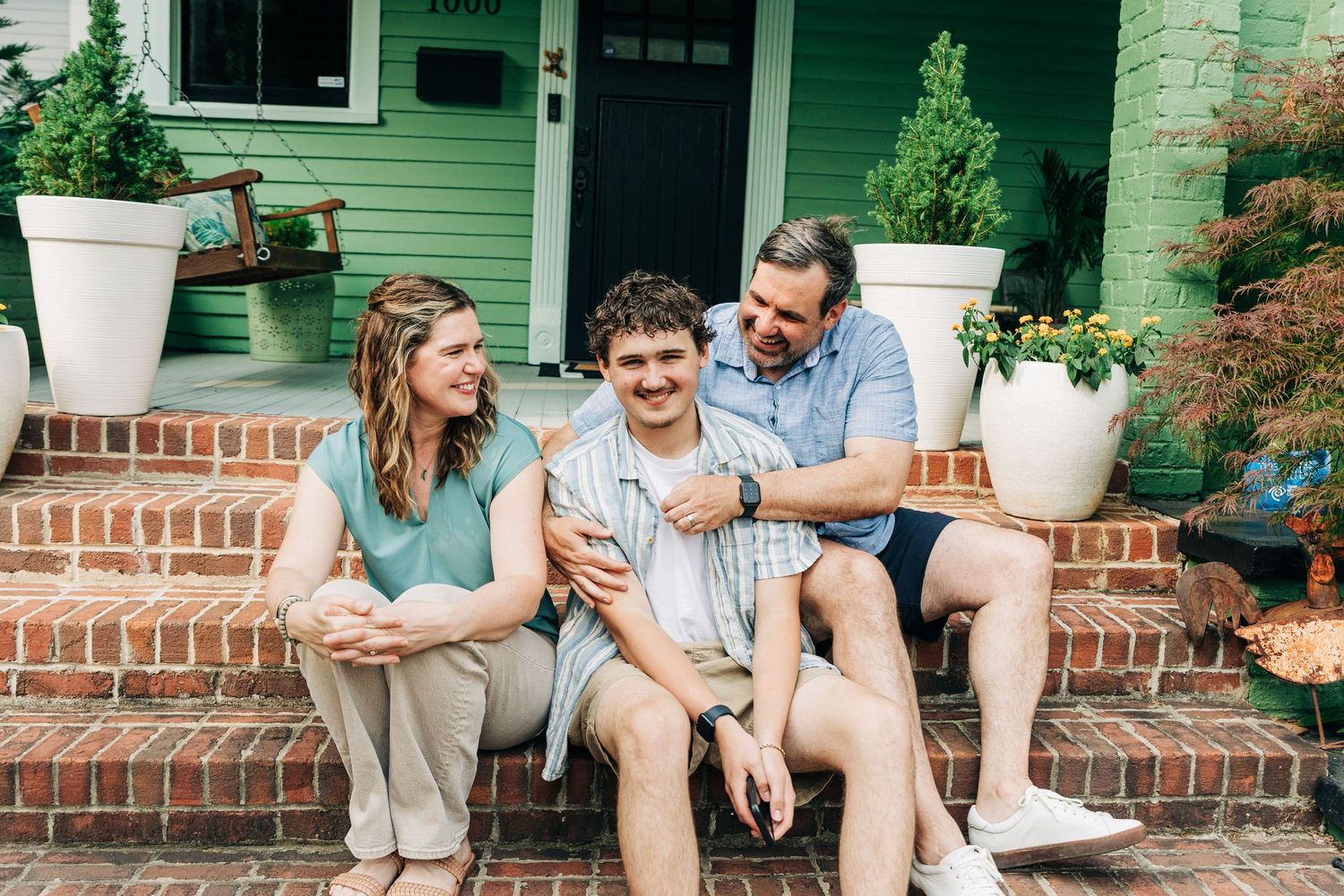 Charlotte family sitting and laughing together on brick steps outside a green house on a sunny day.