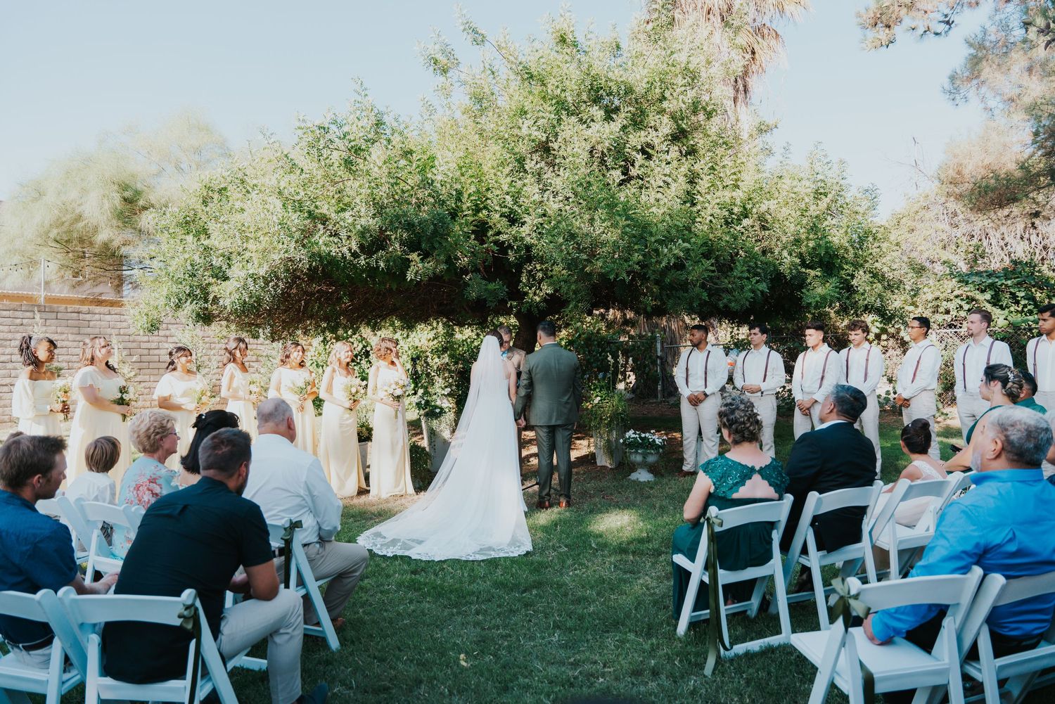 Panoramic view of an outdoor wedding ceremony with guests seated in white chairs on a grassy lawn surrounded by trees.