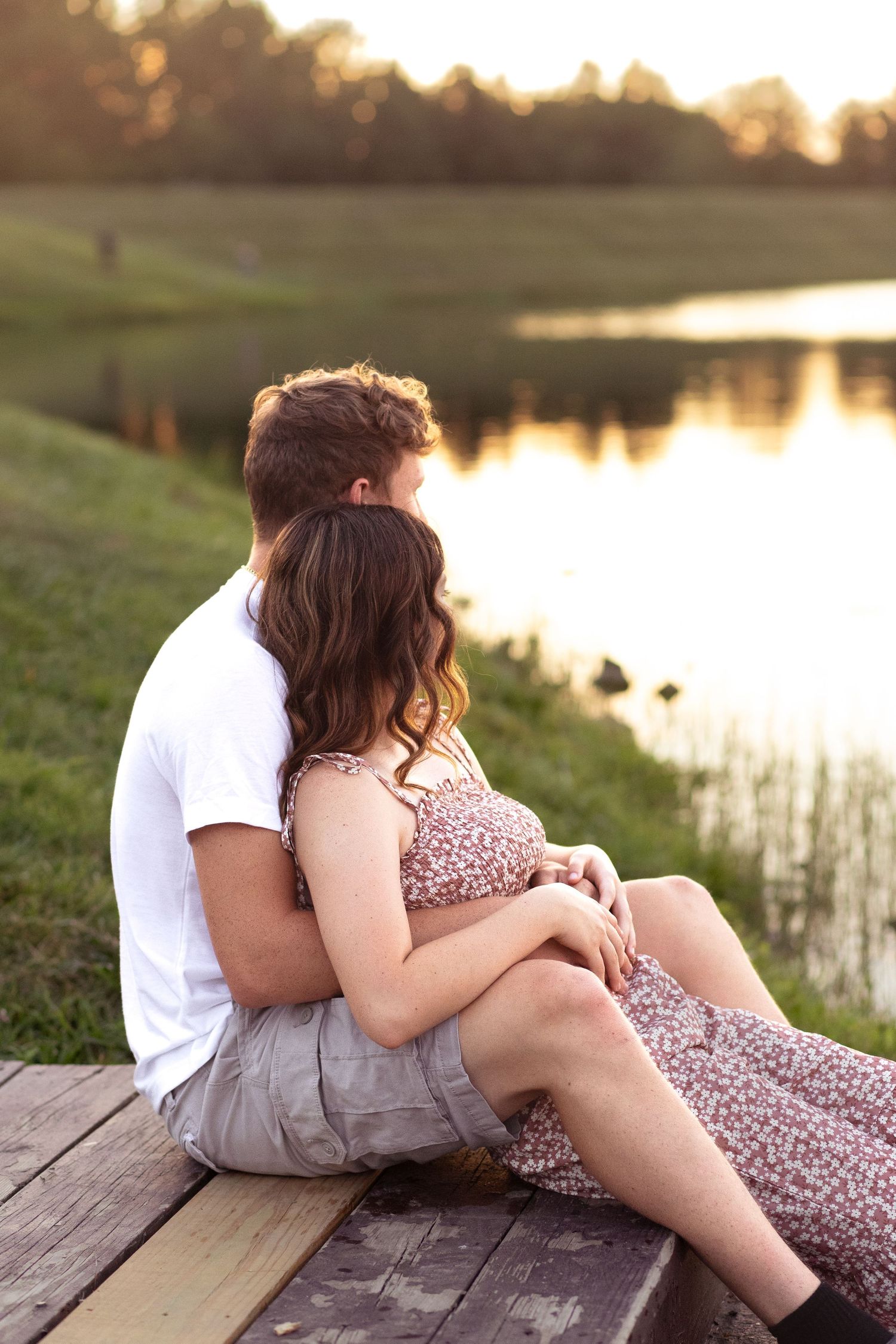 A couple in casual winter attire stands together on a grassy hill overlooking a serene lake on an overcast day.
