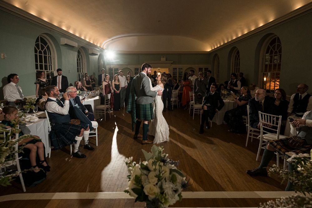 Wedding guests dance together on a wooden dance floor in an elegant reception hall with warm ambient lighting.