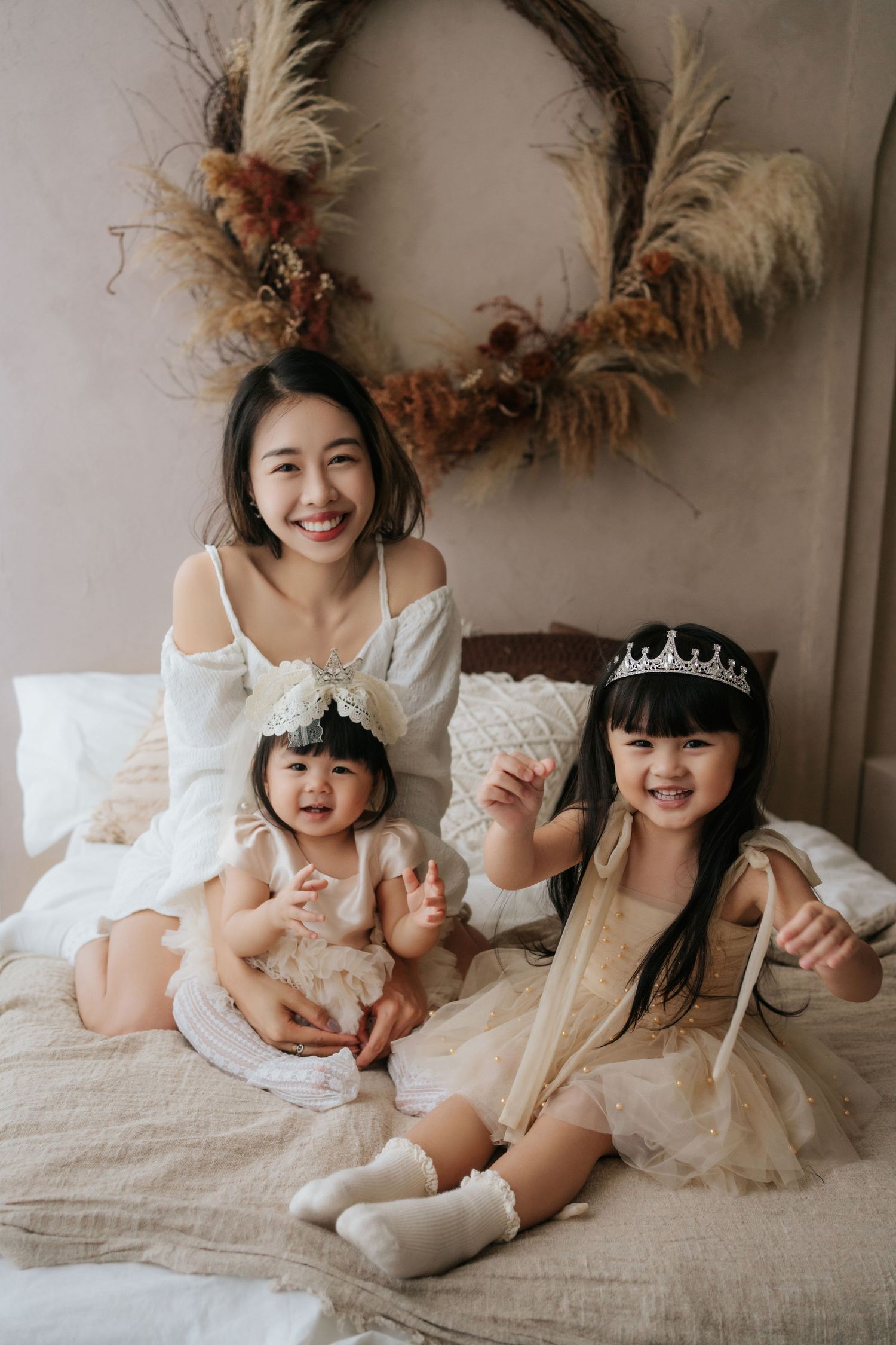 Three people wearing white outfits lay together on a bed with a decorative wreath on the wall behind them.