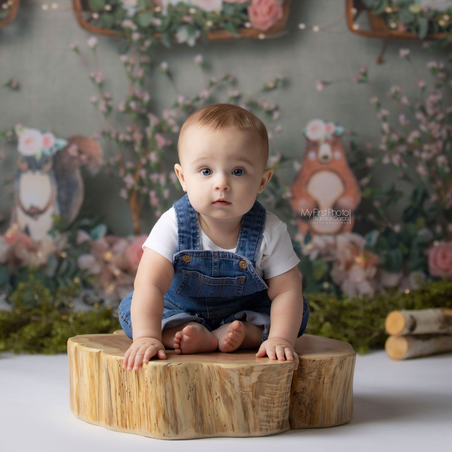 Small child in denim overalls leans over a rustic wood slice on floral backdrop.