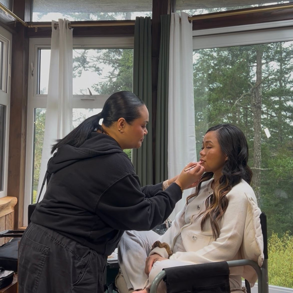 A makeup artist applies cosmetics to a client seated by large windows with forest views in the background.