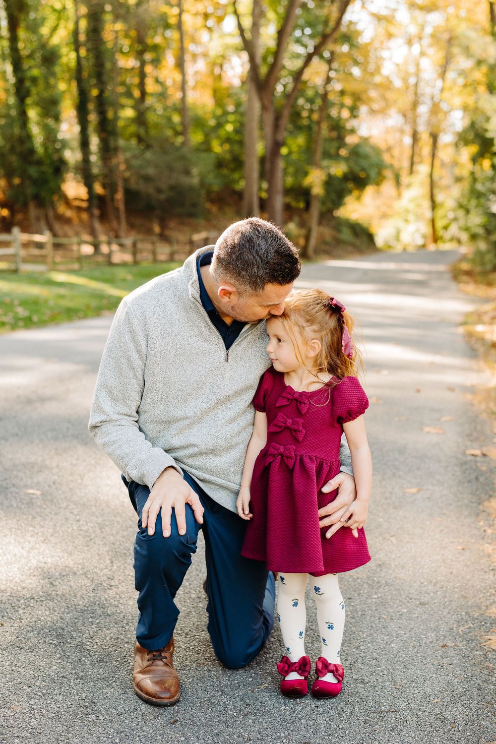 Parent and child share a tender moment while kneeling on a tree-lined path in autumn.