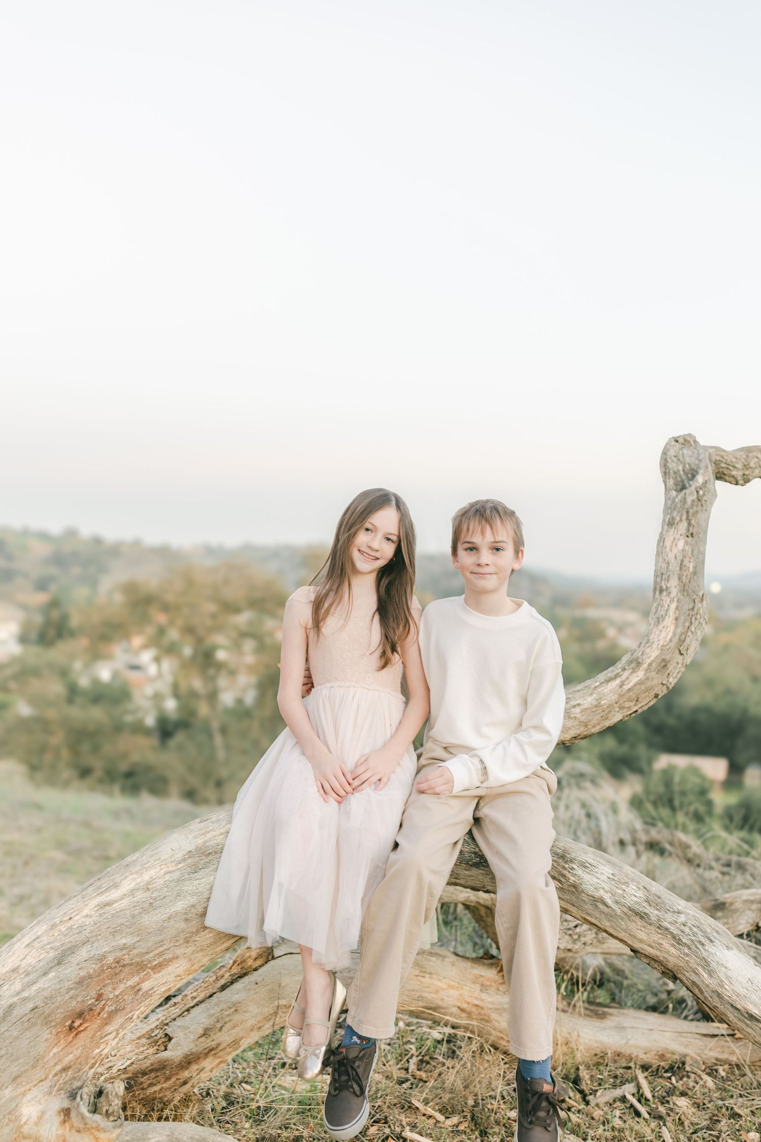 Two young people in light colored clothing sit together on a large piece of driftwood on a hilltop at sunset.