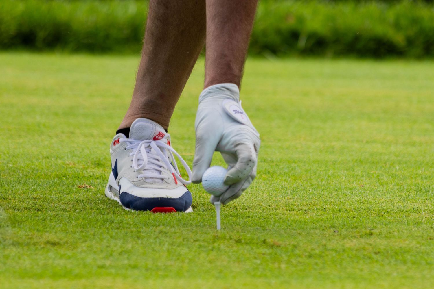 Golfer placing a white golf ball on a tee on a lush green course while wearing a golf glove and white sneakers.