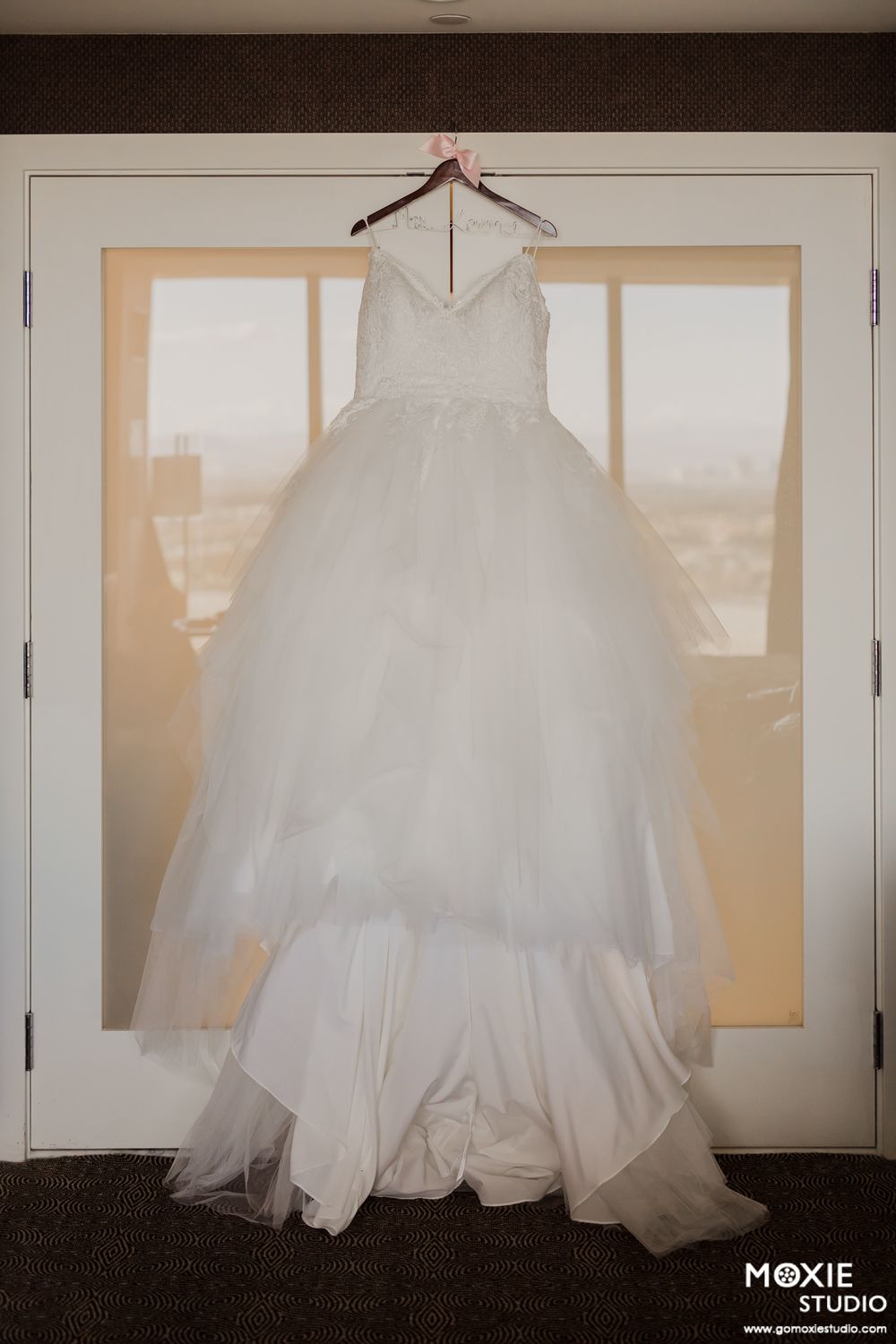 A beautiful white wedding dress hangs elegantly in front of a window, showcasing its full tulle skirt and bodice details.