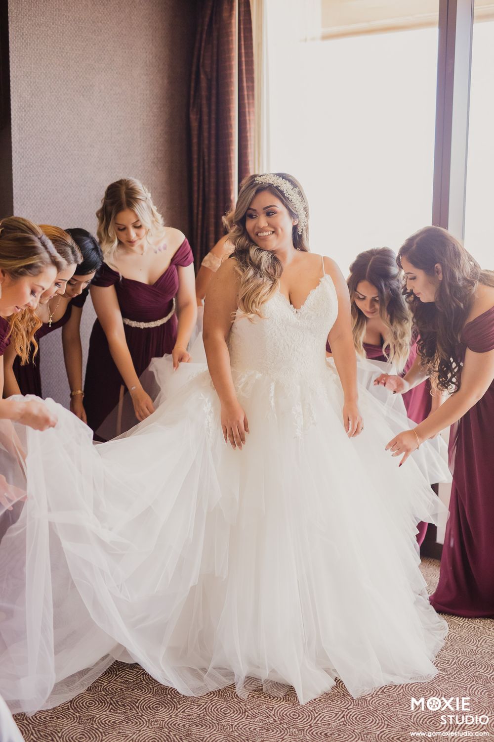 Bridesmaids in burgundy dresses help the bride with her flowing white wedding gown in a sunlit room.