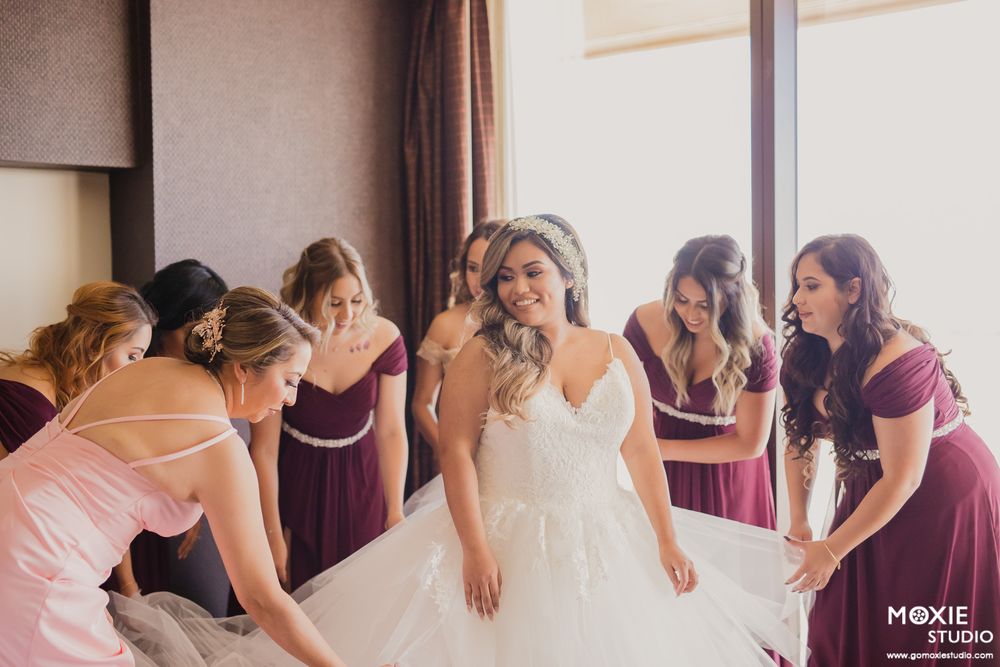 Wedding party adjusts the bride's elegant white dress while getting ready in a hotel room.
