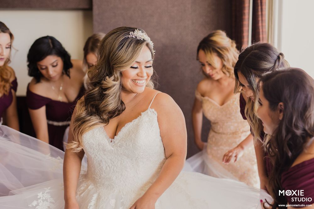 Bridesmaids in soft-colored dresses help with wedding preparations in a sunlit room.