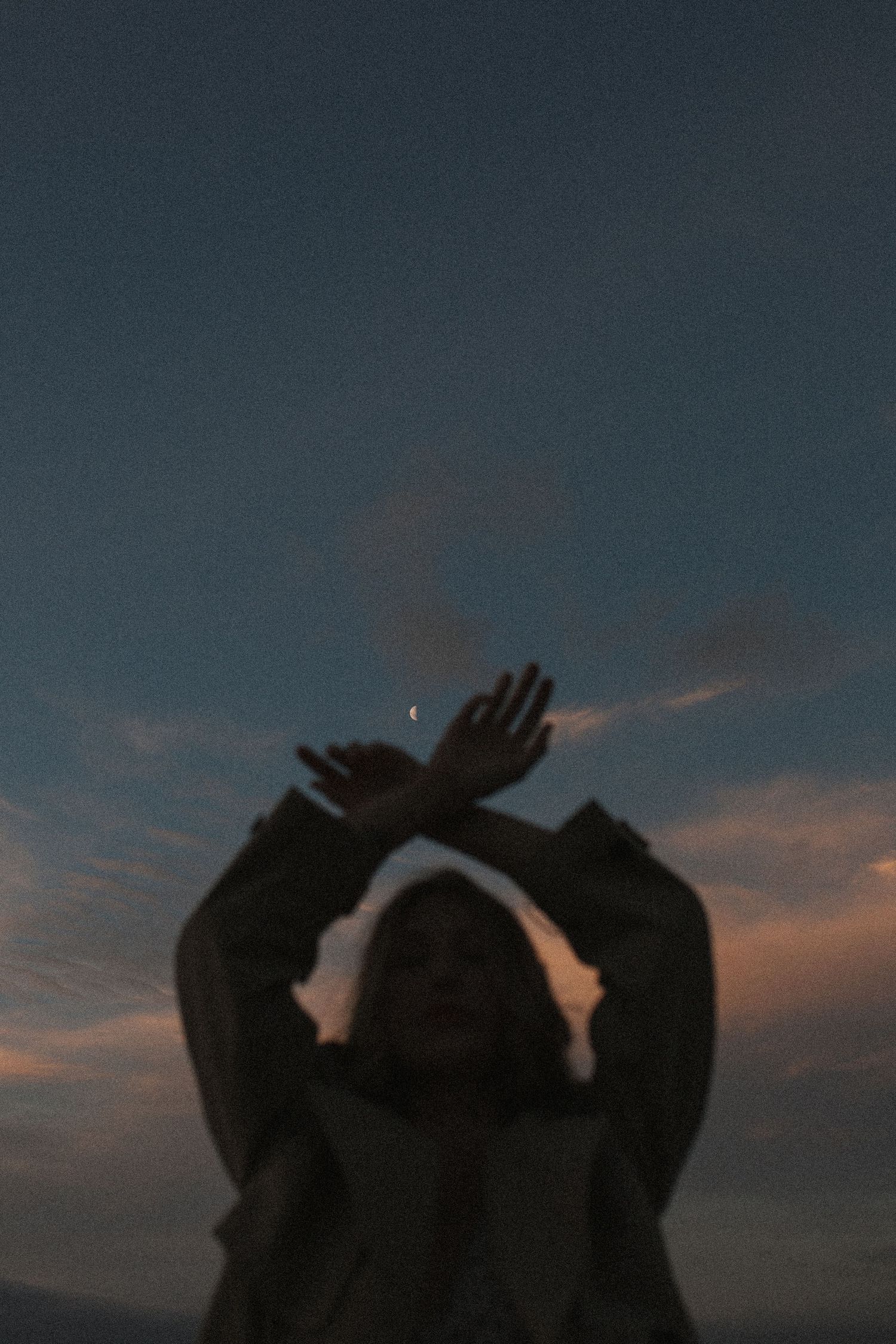 Silhouette of a person raising crossed hands toward a crescent moon during a colorful dusk sky in New Zealand.