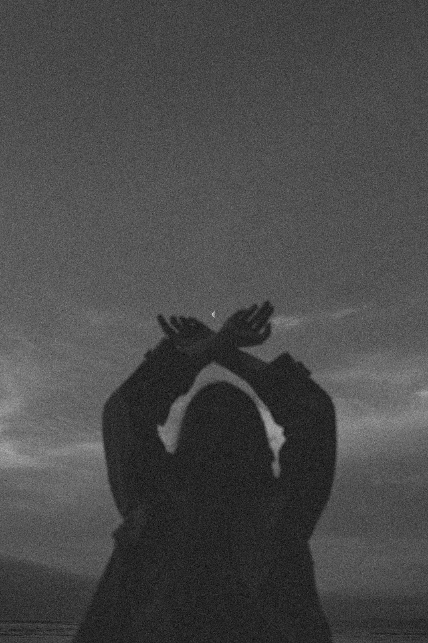 Black and white silhouette of a person raising crossed hands toward a crescent moon in a twilight sky in New Zealand.